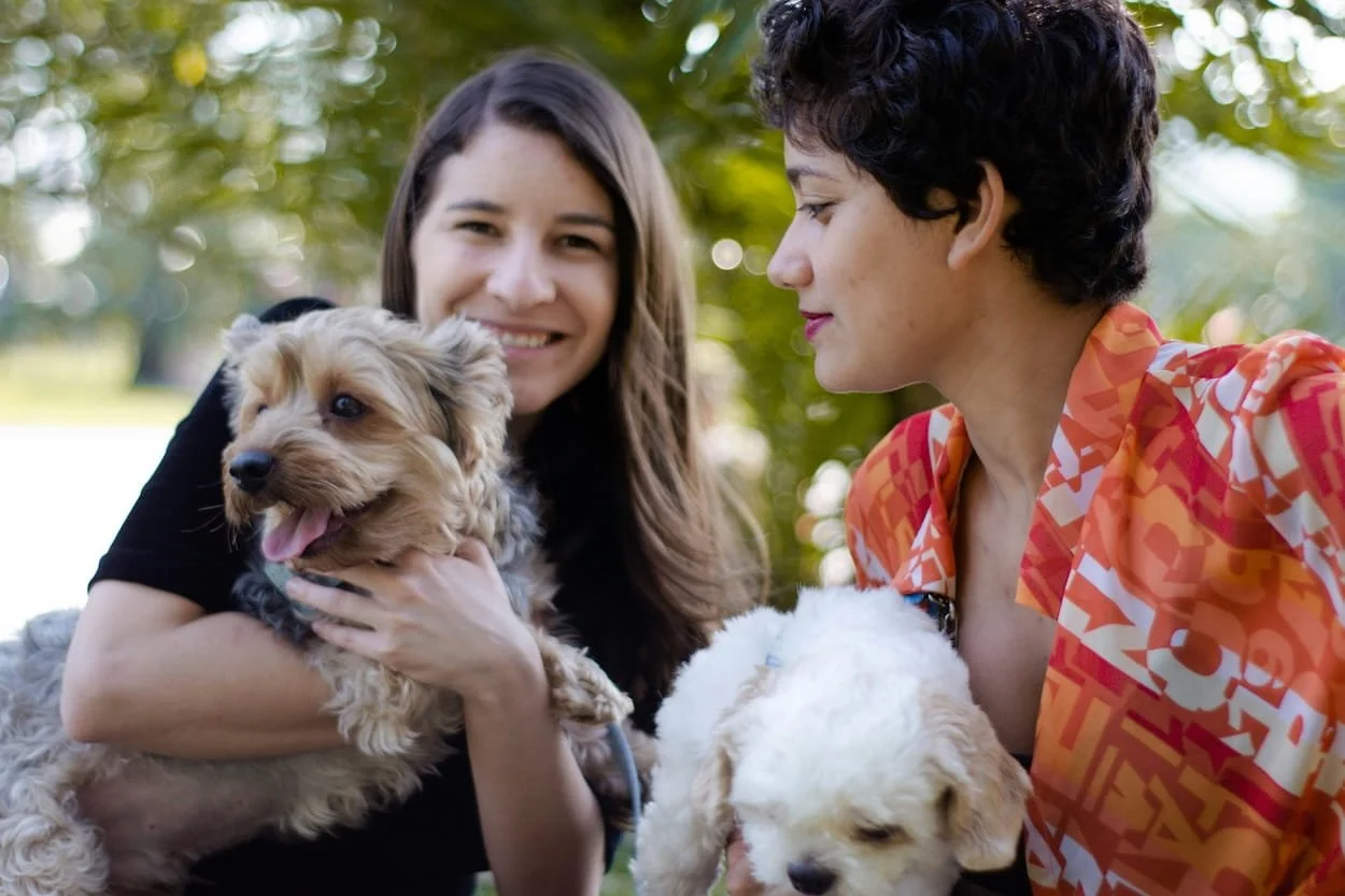 Two women holding dogs in a park with trees in the background, smiling and looking at each other.