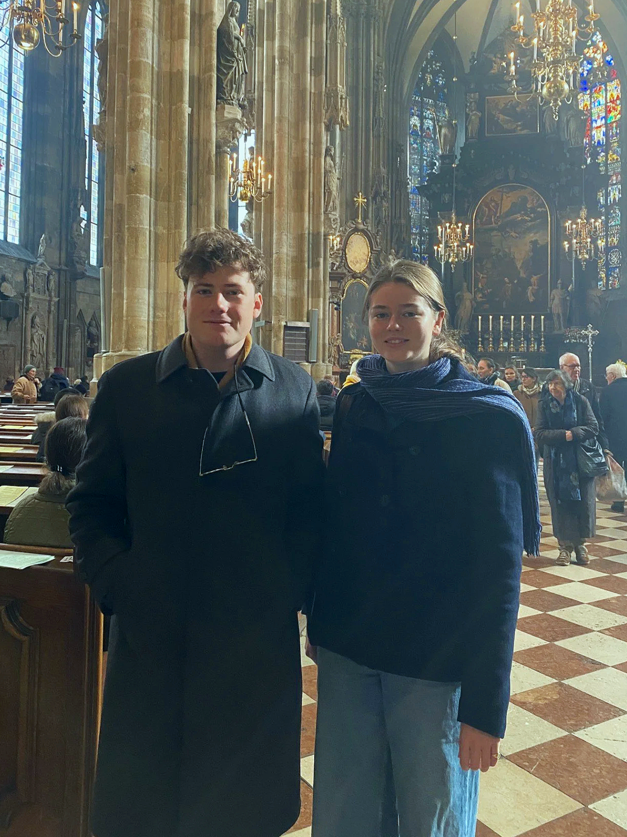 A young man and woman standing inside a historic church with high vaulted ceilings, stained glass windows, and ornate chandeliers. They are dressed casually and are posing for the photo.