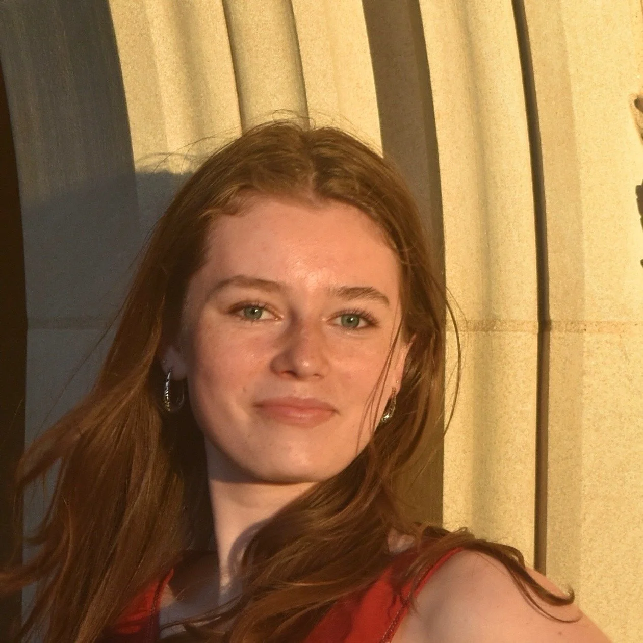 A young woman with red hair, light skin, and green eyes, wearing earrings and a red top, standing near a beige curtain with sunlight illuminating her face.