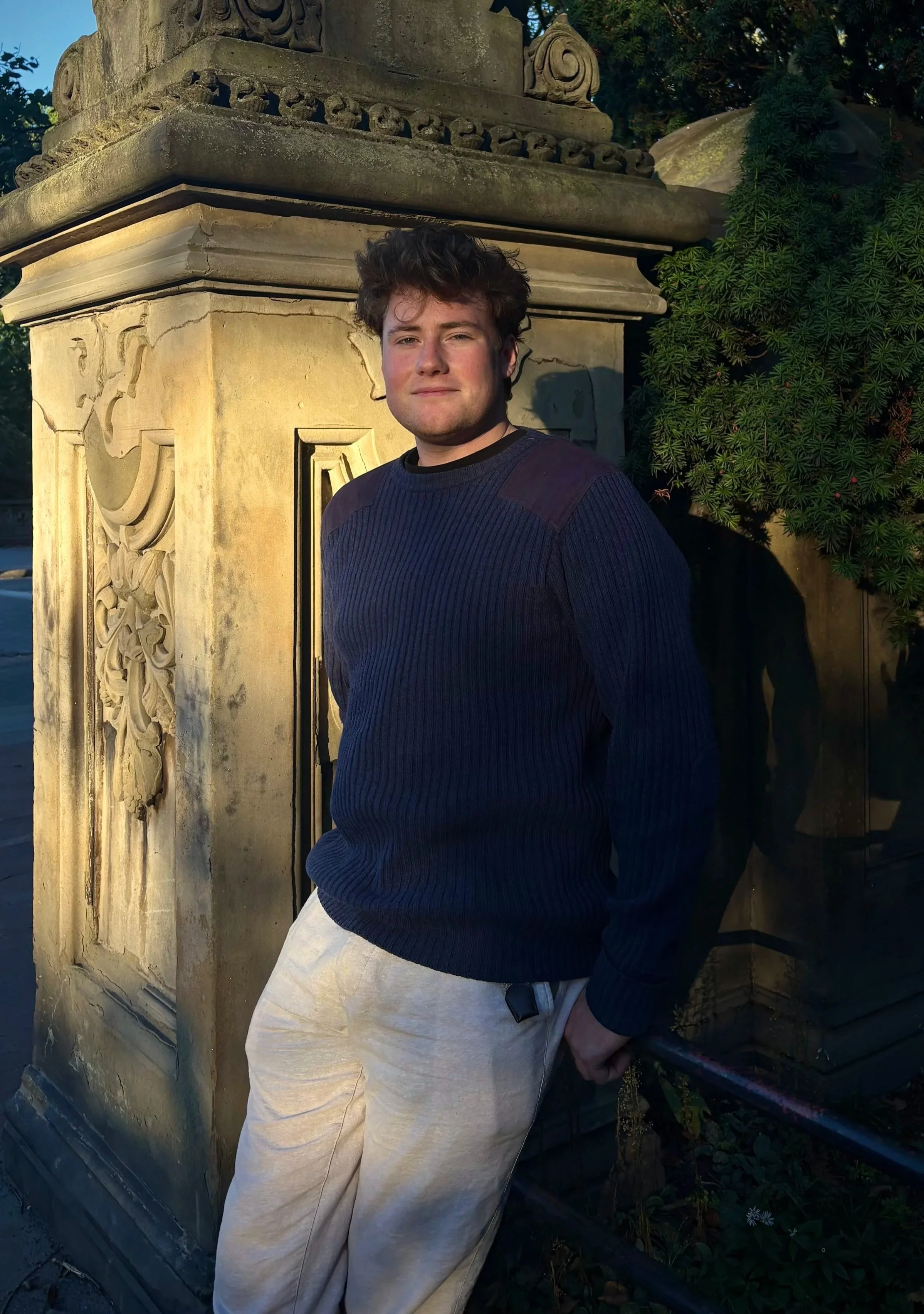 A young man with curly brown hair and light skin, wearing a navy blue sweater and light-colored pants, standing outdoors next to an ornate stone structure with carved designs, illuminated by warm sunlight, and greenery in the background.