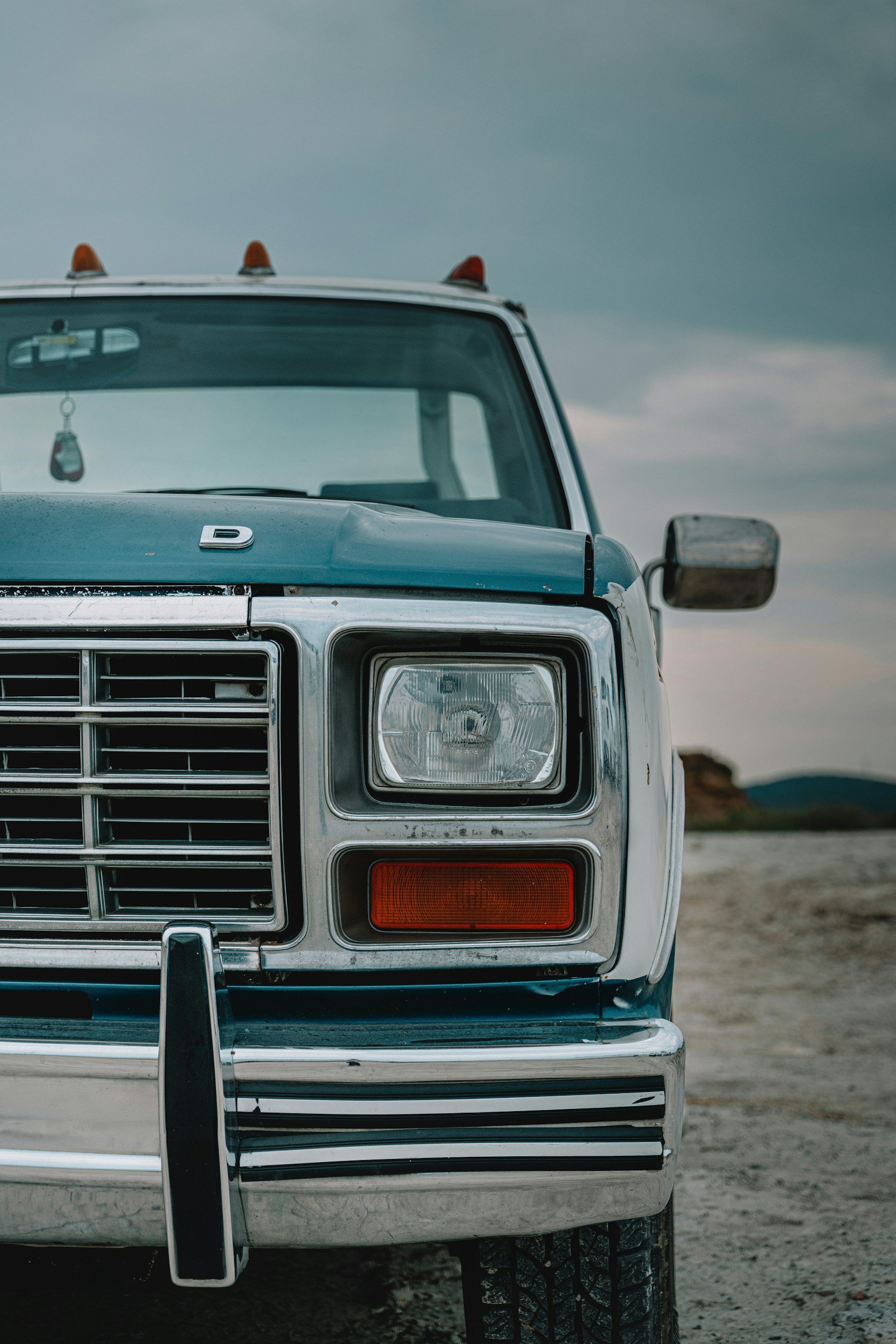 Close-up of the front of a vintage Datsun pickup truck, showing the headlight, grille, and bumper, with an outdoor landscape and cloudy sky in the background.
