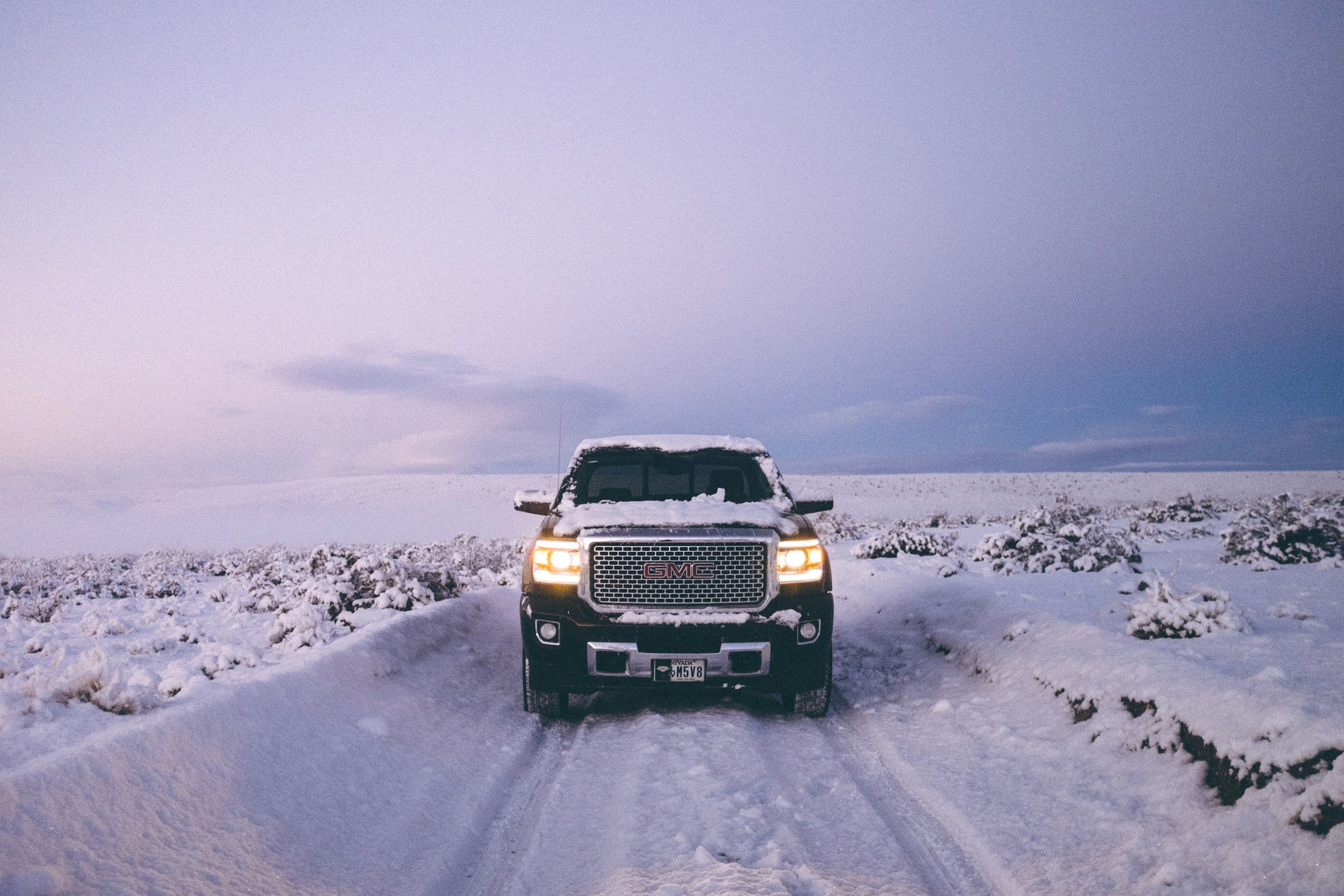 A black GMC pickup truck parked on a snow-covered rural road with snow-covered fields and a cloudy sky in the background.