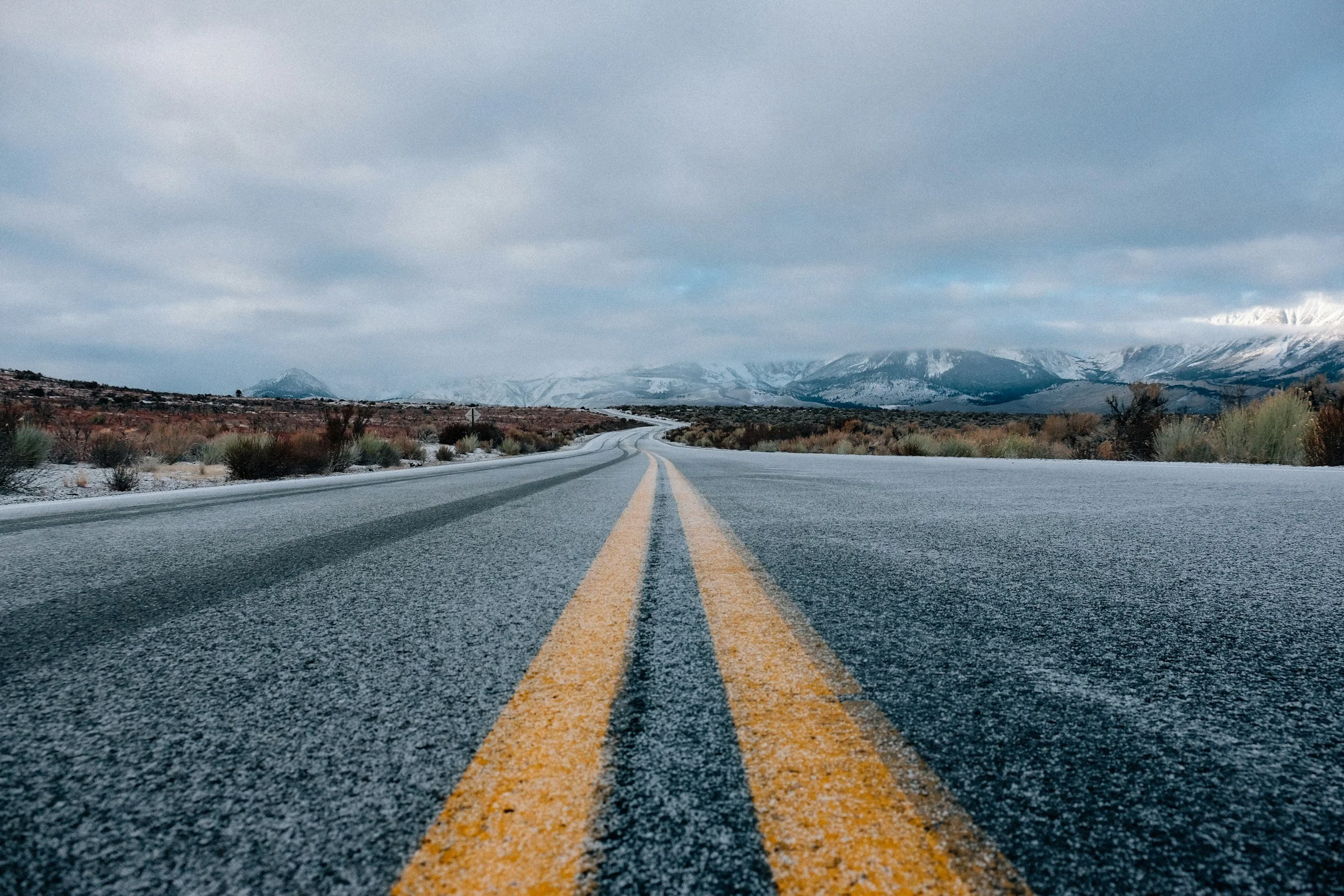 A straight paved road with yellow double lines in a winter landscape, with snow on the ground and mountains in the distance under cloudy sky.