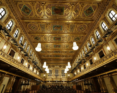 The interior of an ornate, historic theater or opera house with a decorated ceiling featuring gold accents and painted murals, multiple balconies with intricate railings, and large hanging chandeliers.