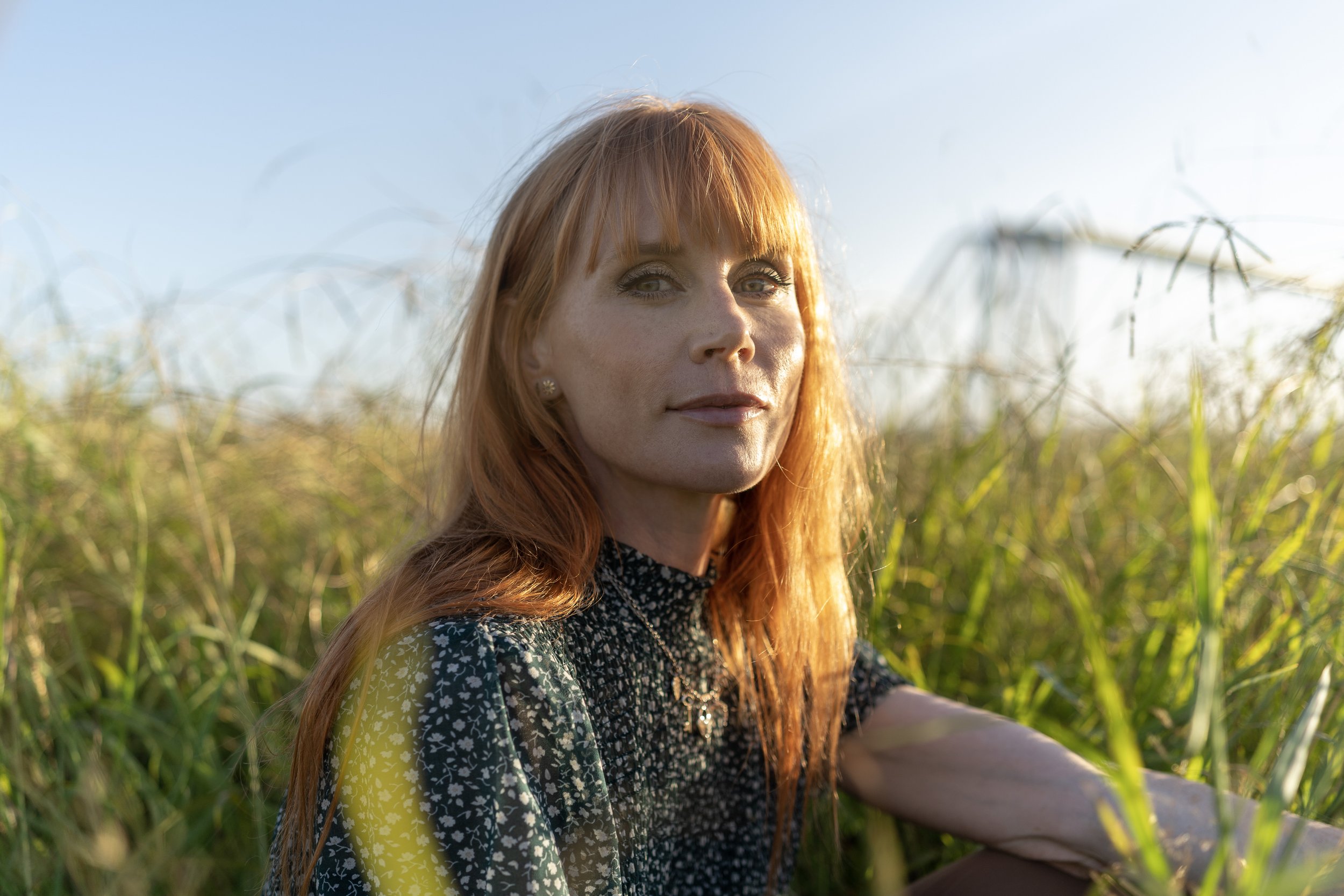 New Zealand actor Michelle Blundell photographed by Emma Cosgrave. She is looking to camera with a warm smile and long red hair sitting in long green grass