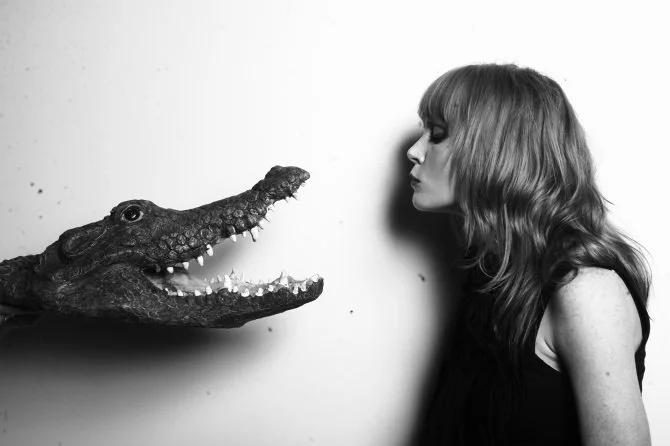 A black and white photograph of actor Michelle Blundell taken by Michelle Hyslop, with art direction from Kim Band. Michelle is wearing black singlet and is looking into the open mouth of a fake crocodile