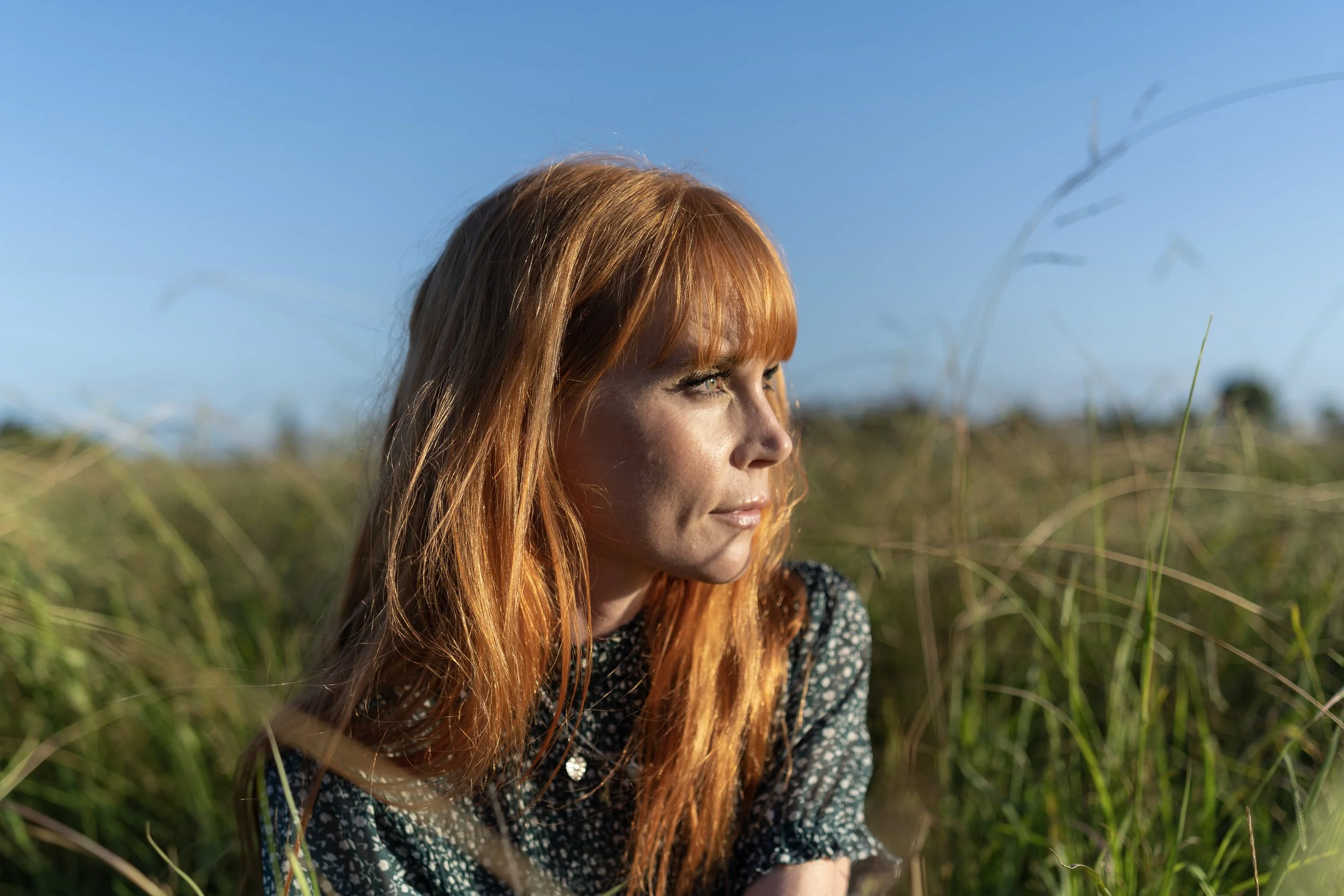New Zealand actor Michelle Blundell photographed by Emma Cosgrave. She is looking away from camera with a warm smile and long red hair sitting in long green grass