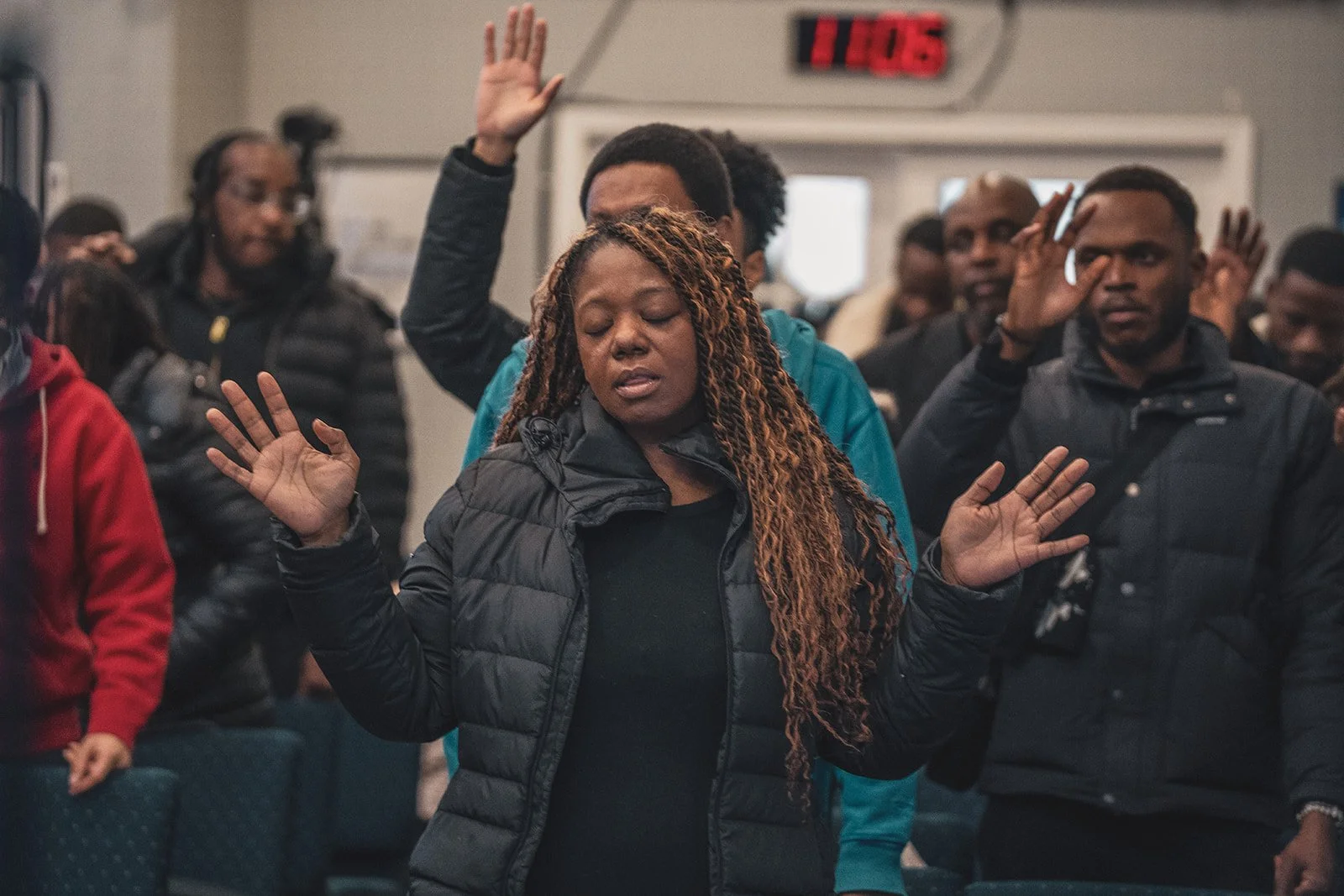 A woman with long braided hair wearing a black puffy jacket praying with her eyes closed and hands raised, surrounded by other people in a church or prayer meeting.