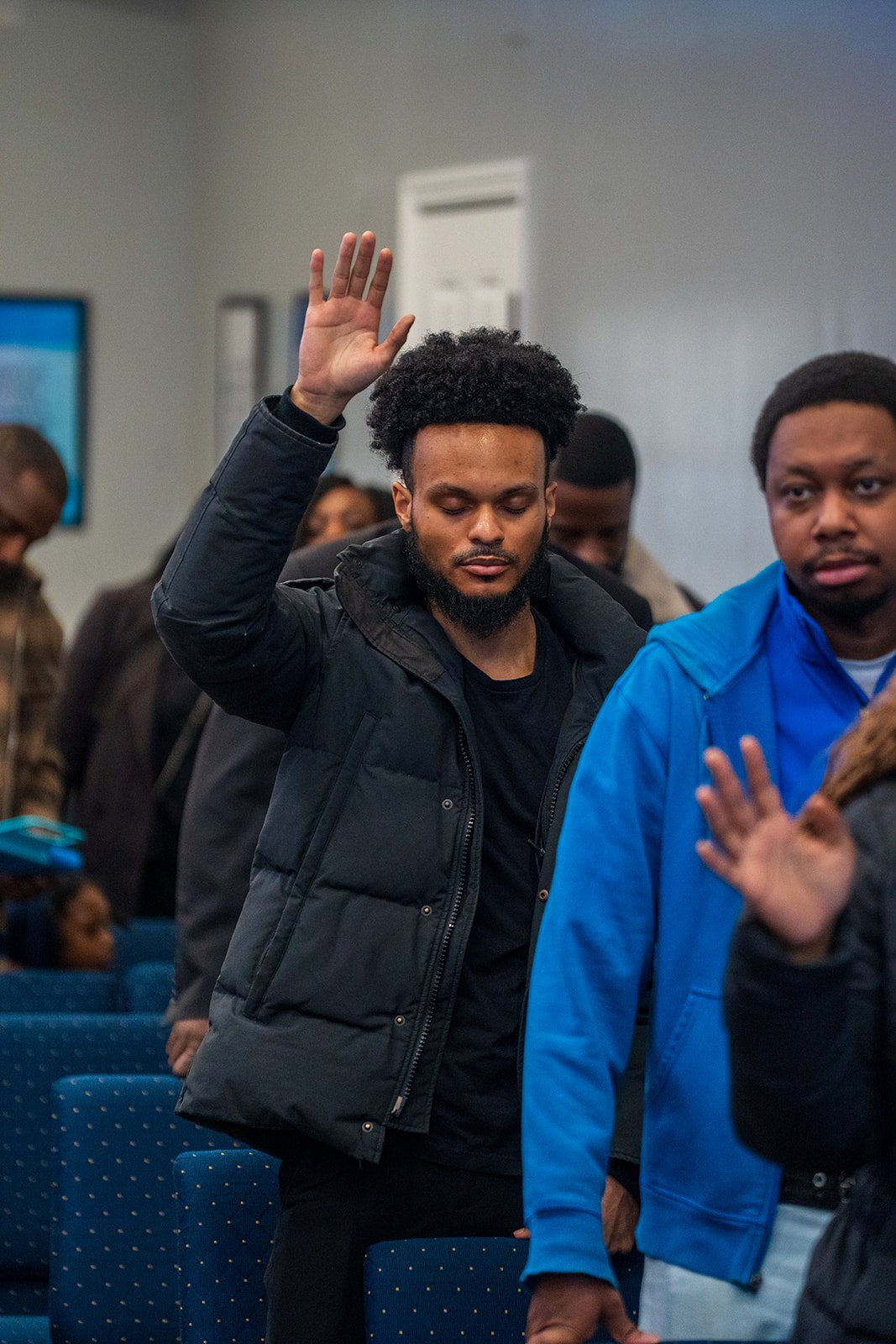 A man with curly hair and a beard raises his hand during a meeting or event, standing among other people in a room with blue chairs.