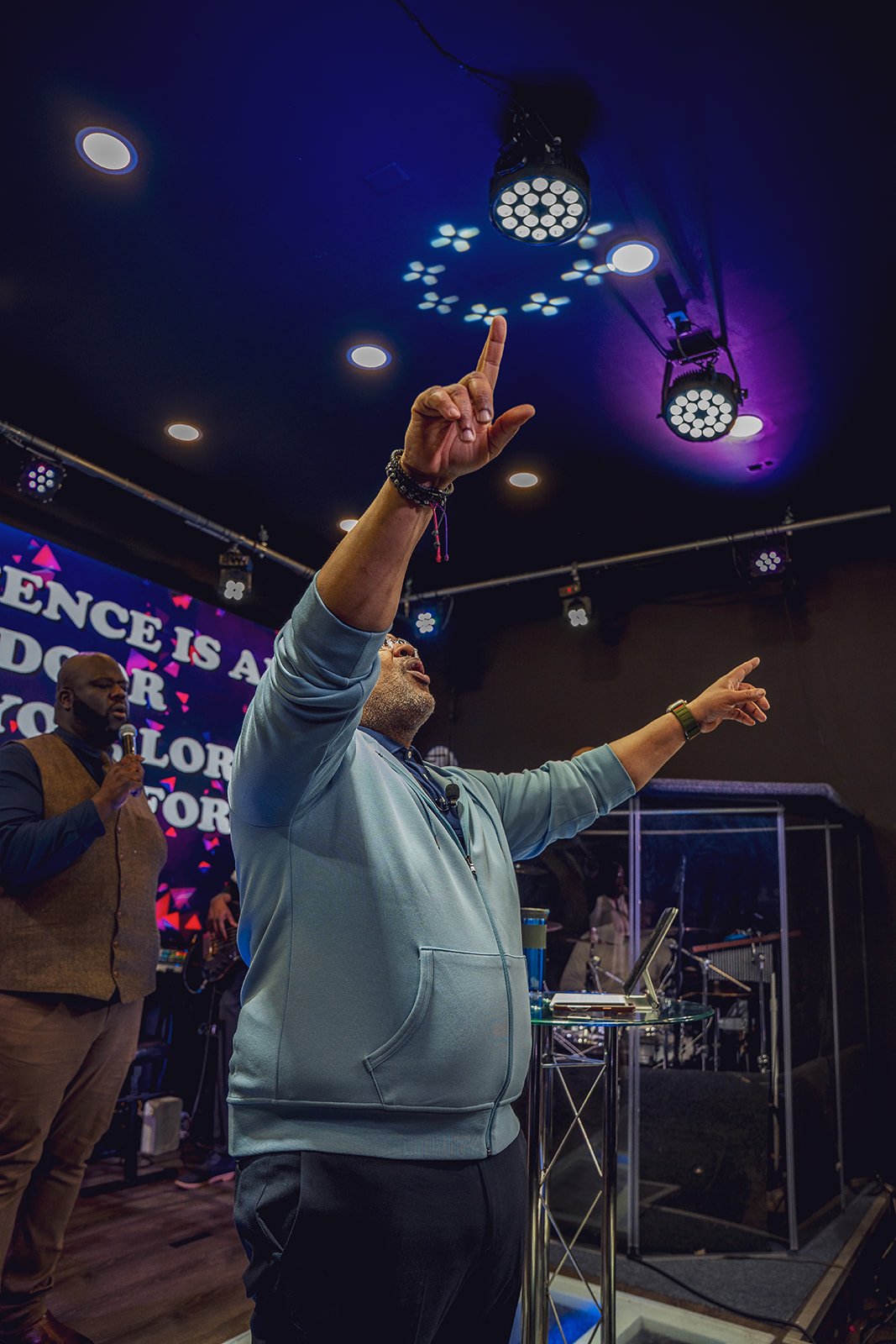 A man in a light blue hoodie with glasses praying or worshipping with one hand raised and finger pointing upward at a church or conference. Other individuals are present, and a large screen with colorful text and stage lighting are visible in the bac