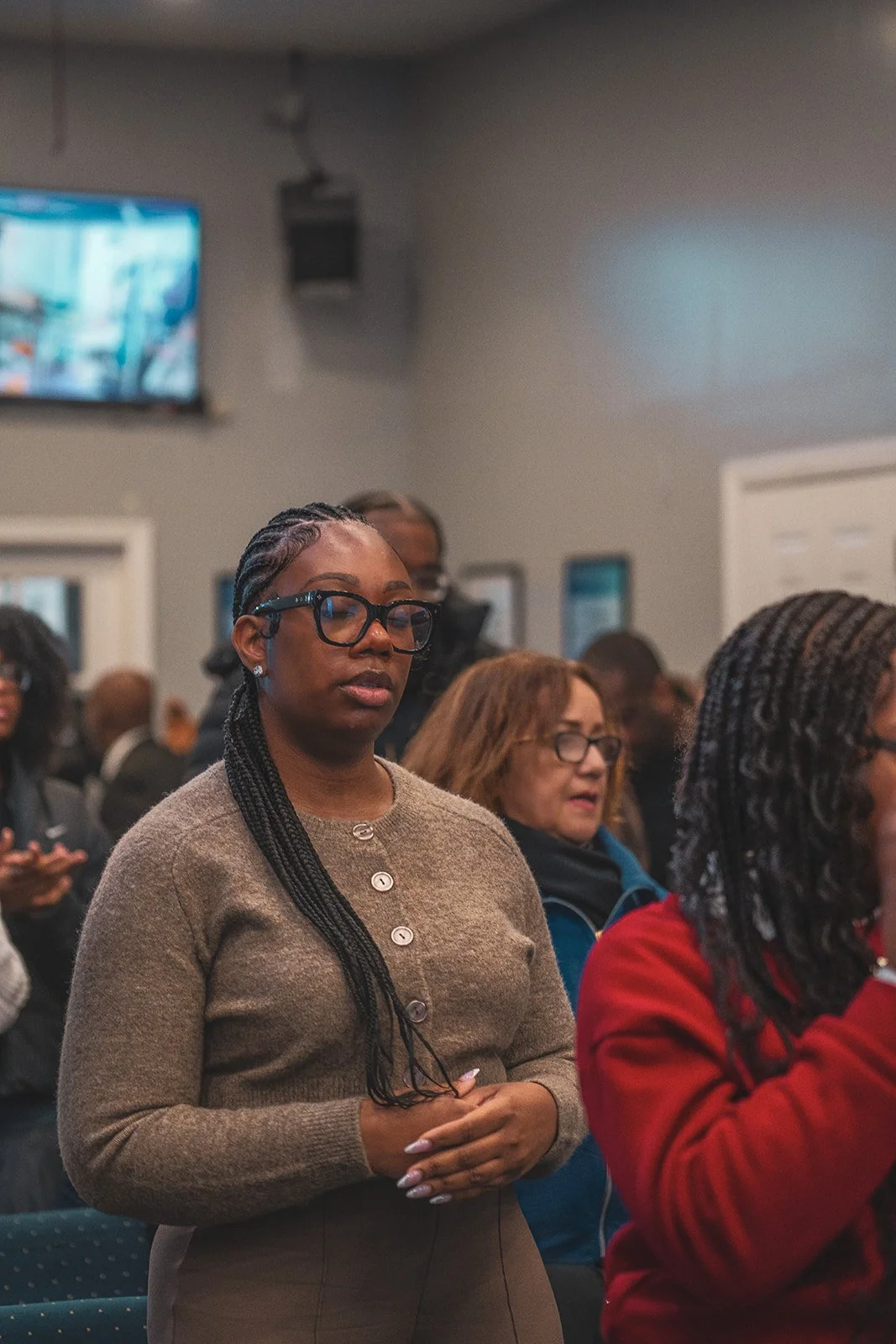A woman with braided hair, glasses, and a beige sweater standing with her eyes closed in a room full of people attending an event.