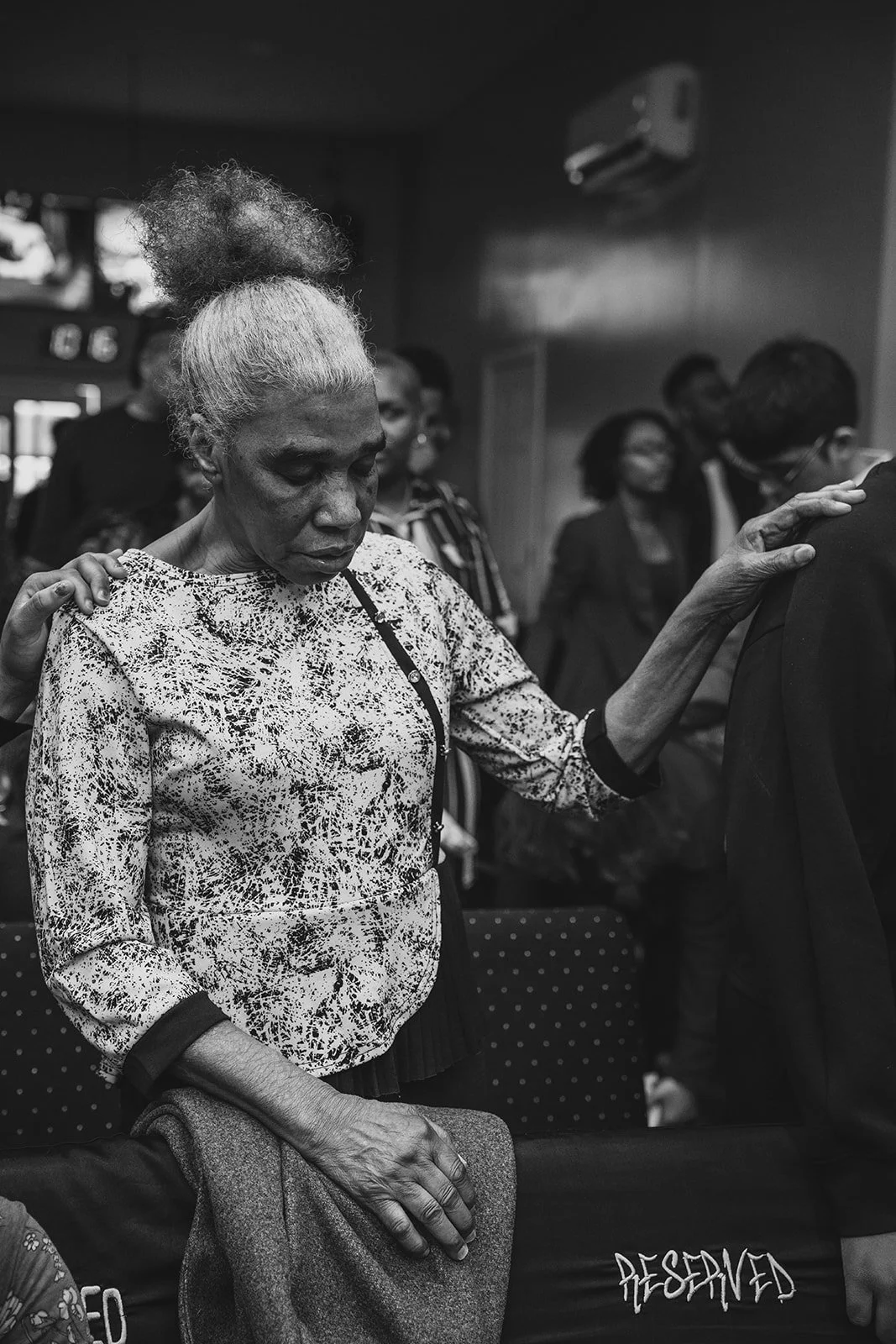 An elderly woman with gray hair in a high puff is praying with her eyes closed in a church, with her hand resting on a man's shoulder.