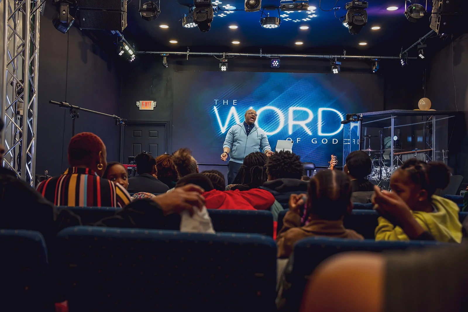 A man on stage in front of a large screen that says 'The Word of God' in a darkened room with audience members seated and listening.