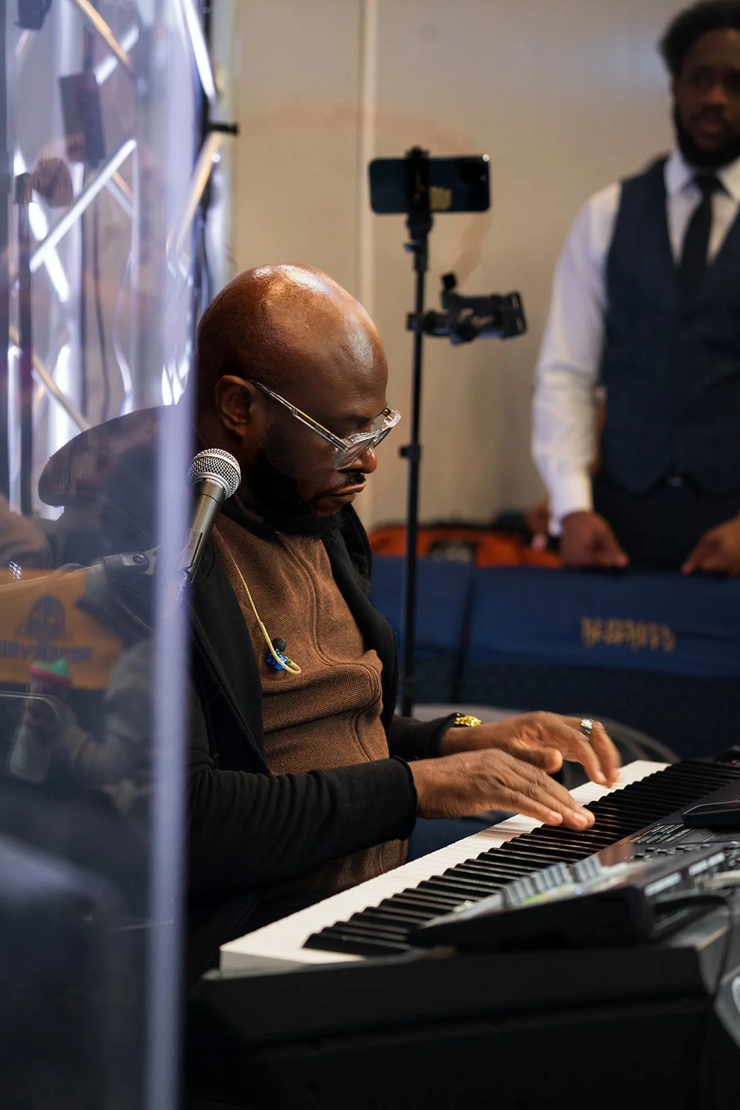 A man playing the keyboard at a music event, with a person standing in the background. The musician is wearing glasses and a brown shirt, focused on playing.