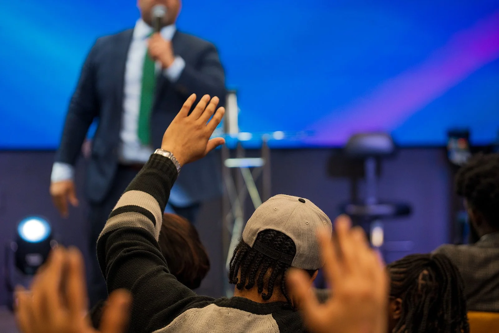 Audience members raise their hands during a presentation or conference, with a man in a suit speaking on stage in the background.