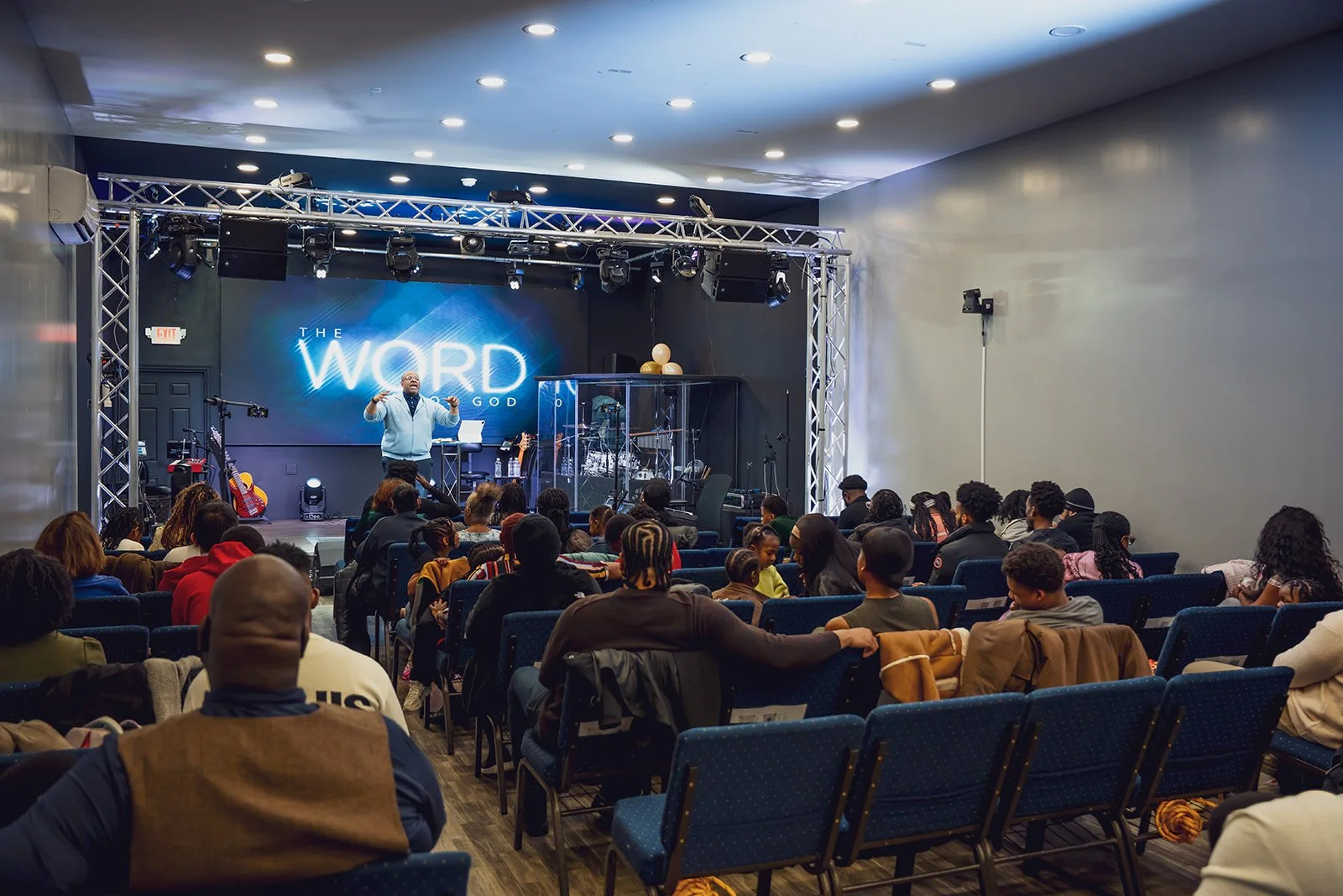 A speaker on stage presenting to an audience in a church or event hall. The background displays a large screen with the text 'The Word of God.' Audience members sit in blue chairs, some with their arms around each other.