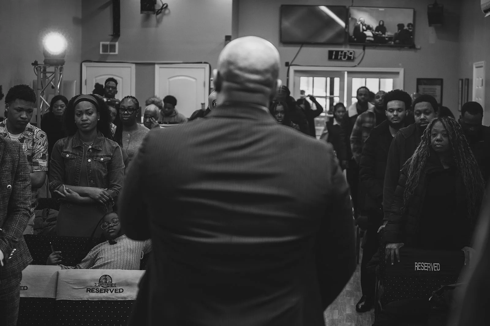 A man with a bald head seen from behind speaking to a diverse group of people in a room, with some chairs labeled 'RESERVED' and a TV screen showing a panel discussion. The group is attentively listening.