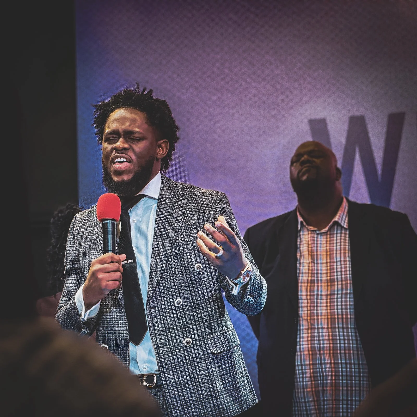 A man in a checkered blazer speaking passionately into a red microphone on stage, with another man in a suit and plaid shirt in the background.