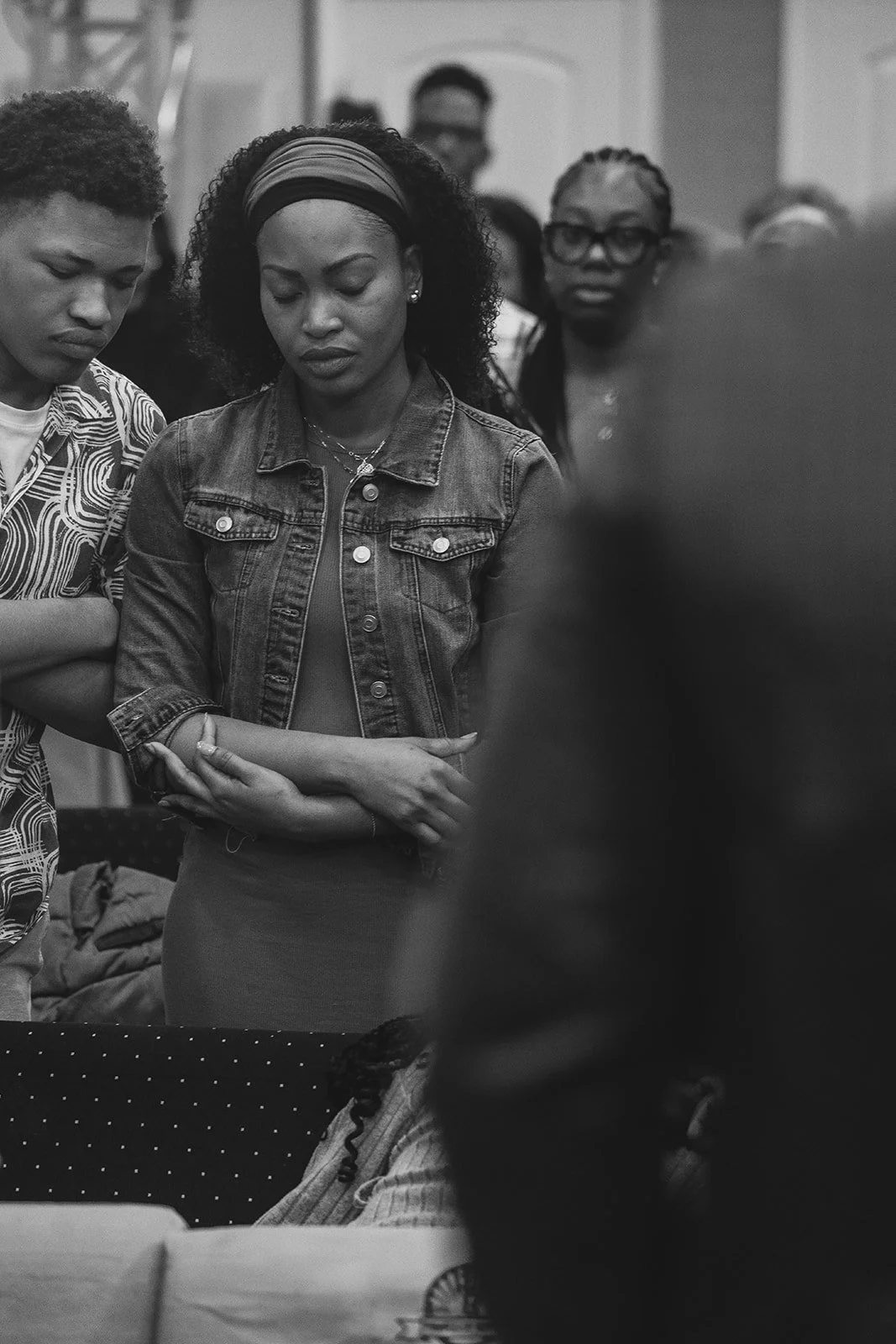 A group of people gathered indoors with one woman at the center, eyes closed, holding her arms crossed, appearing to be in prayer or reflection.