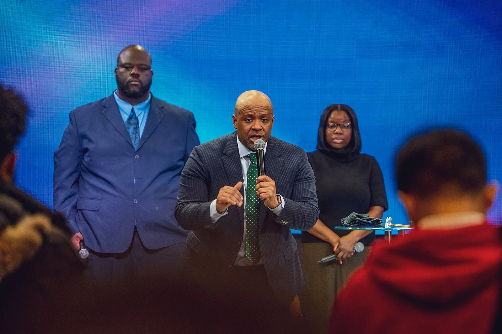 A man in a striped suit and green tie speaks into a microphone at a public event, flanked by two people standing behind him, one man in a blue suit and a woman in black, with an audience in the foreground.