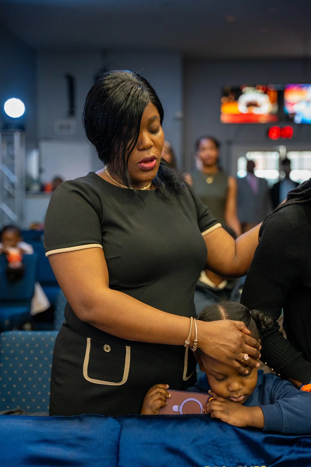 A woman with black hair and a black dress with beige trim gently prays over a young girl with braided hair sitting at a table in a public setting, likely a church or community hall, with other people in the background.