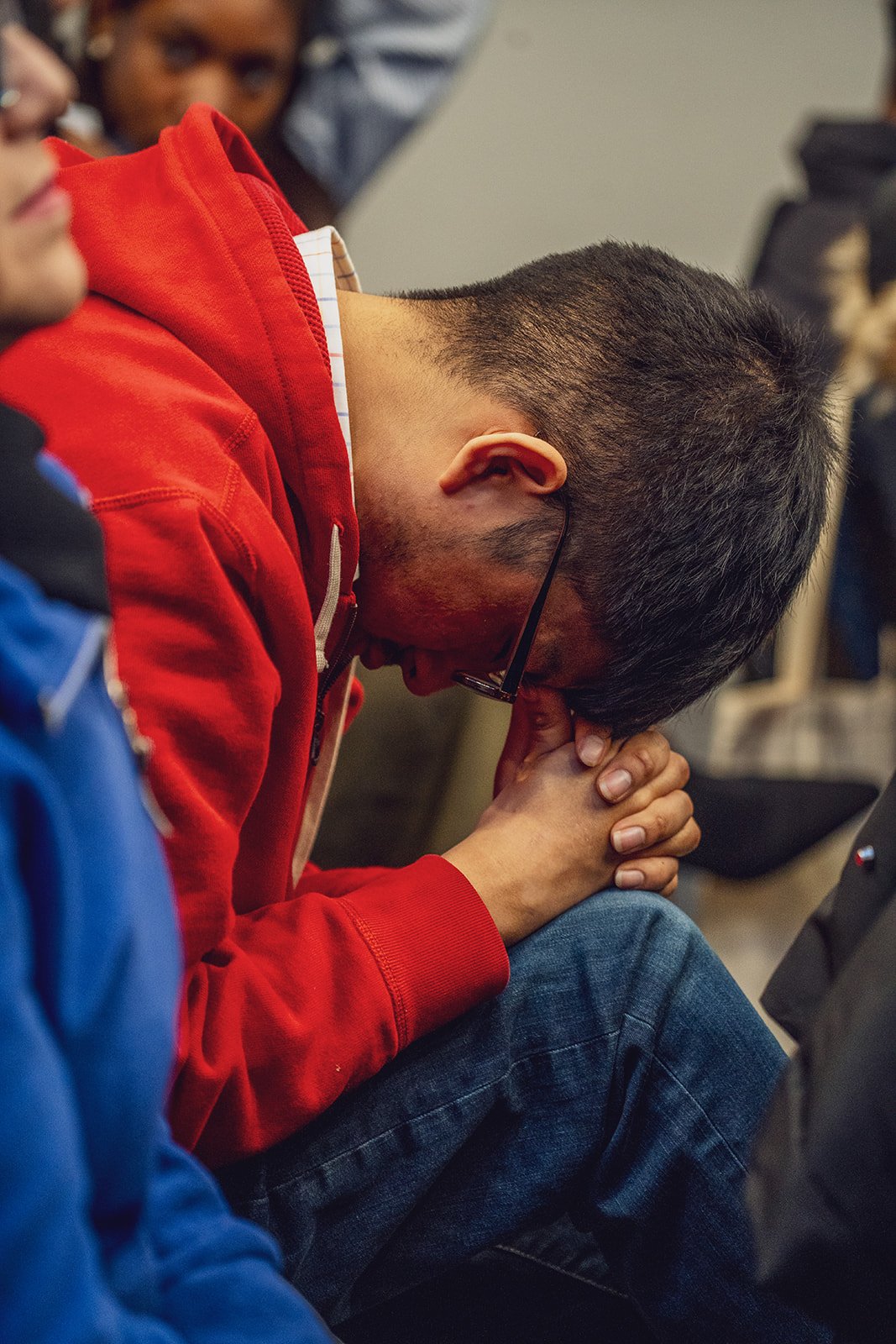 A man with glasses in a red hoodie and dark hair praying with hands clasped and head bowed, surrounded by other people in an indoor setting.