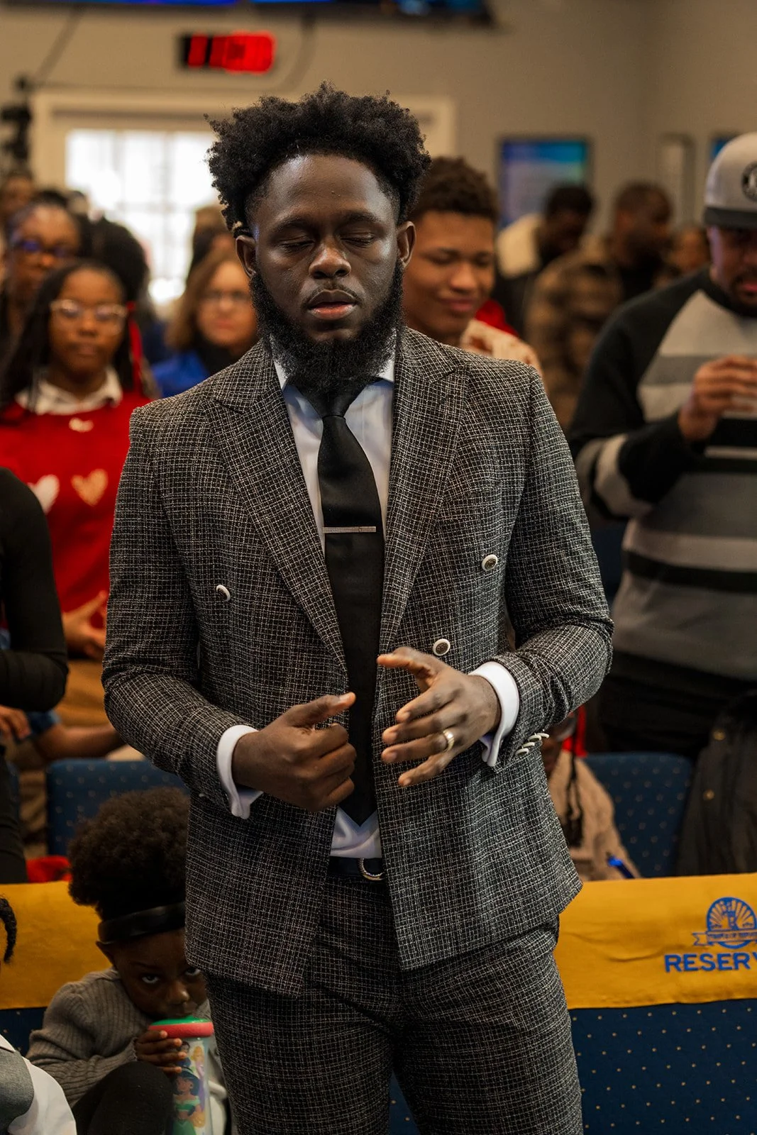 A man with closed eyes wearing a suit and tie, standing with hands clasped, surrounded by an audience in a room.