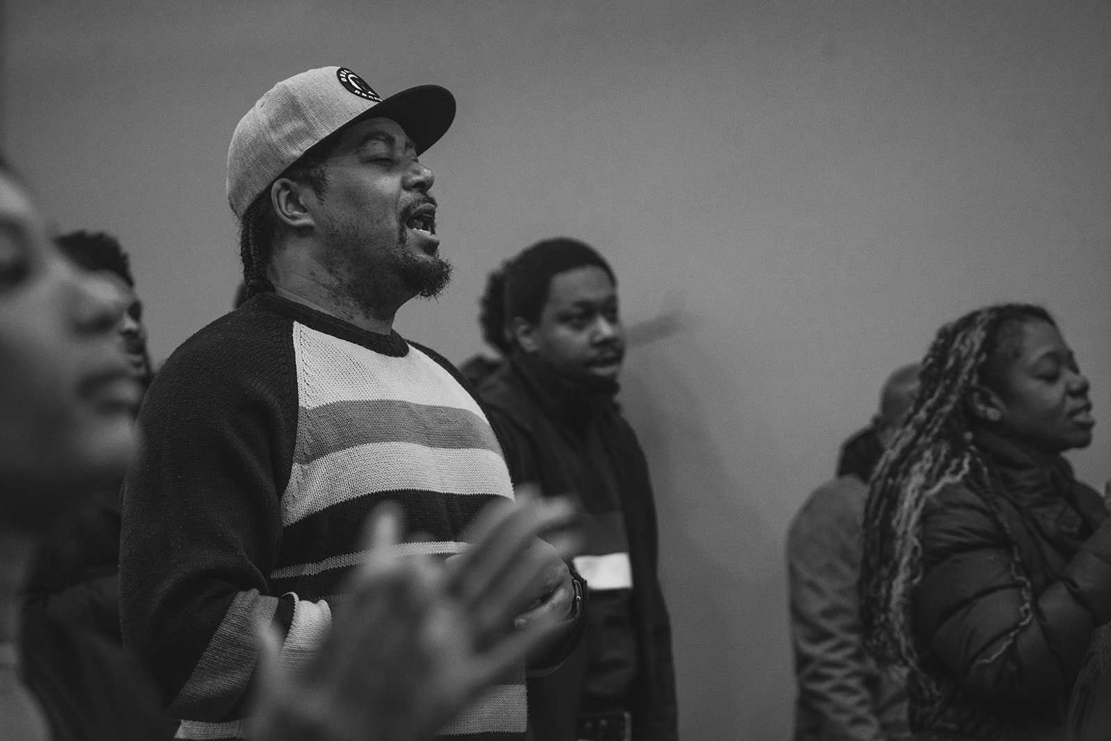 A group of people standing with eyes closed, participating in a prayer or meditation session. The focus is on a man wearing a baseball cap and a striped sweater, with other individuals visible around him.