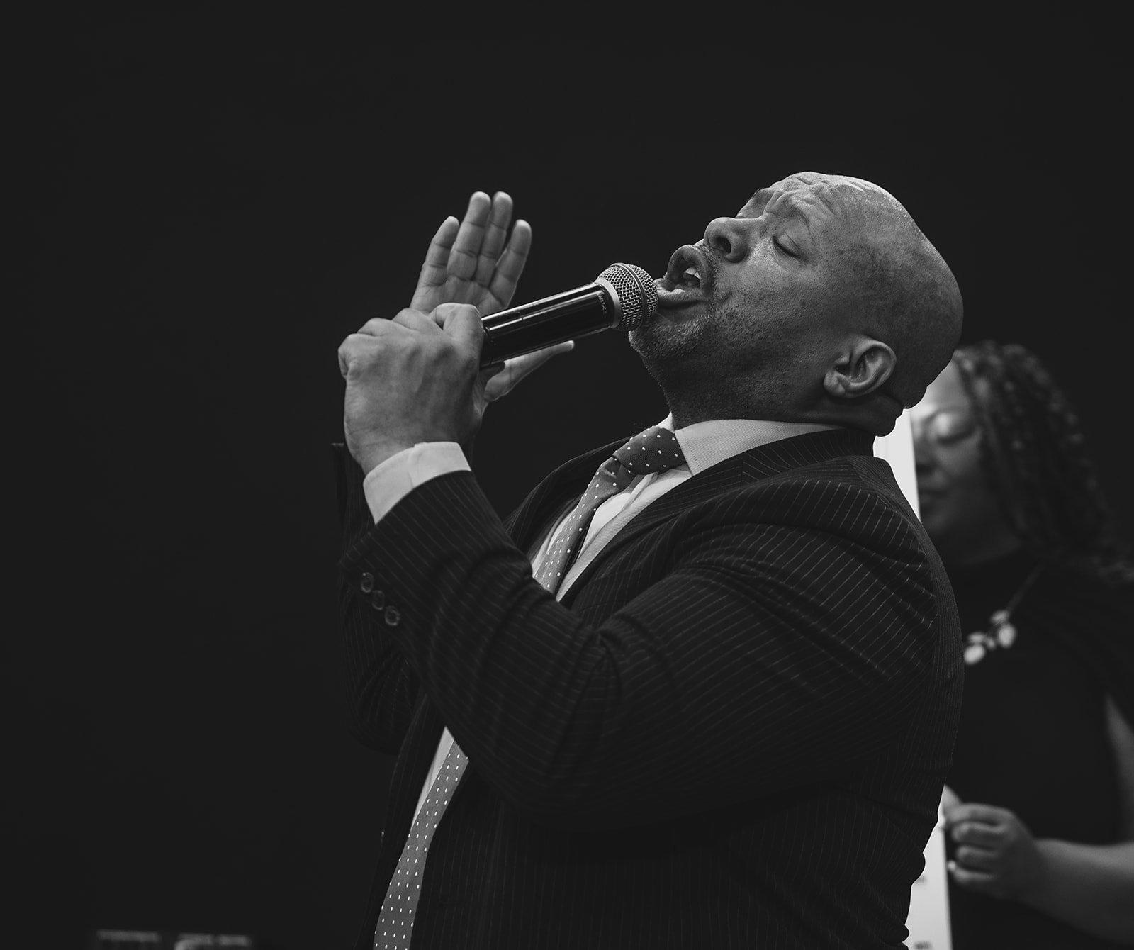 A man in a pinstripe suit, white shirt, and polka dot tie, singing passionately into a microphone, with eyes closed and one hand raised, in a black-and-white photograph.