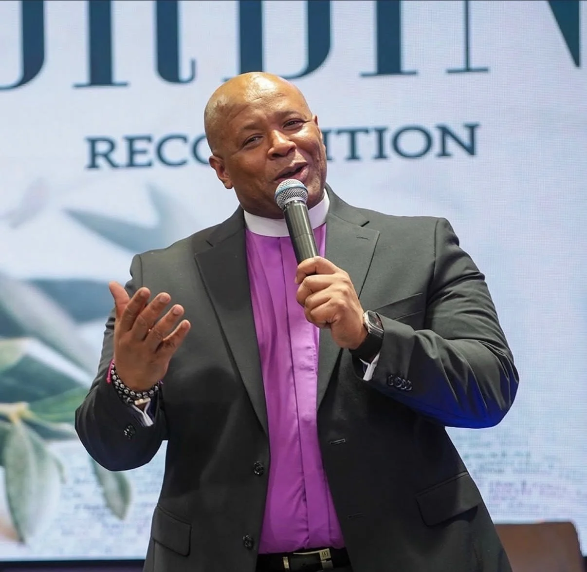 A man in a gray suit and pink clerical shirt speaking into a microphone at an event.
