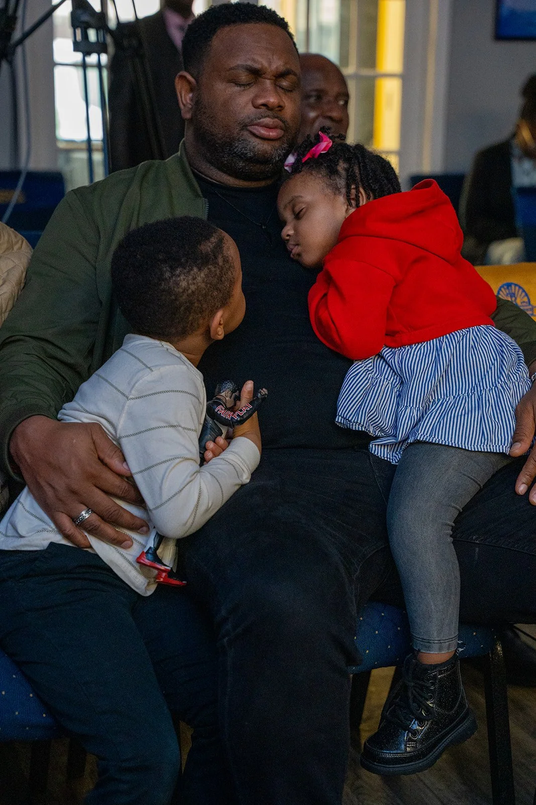 A man with closed eyes sitting with two children resting on him, one girl in a red hoodie with a pink bow in her hair asleep on his chest, a boy in a white striped shirt leaning in, in an indoor setting.