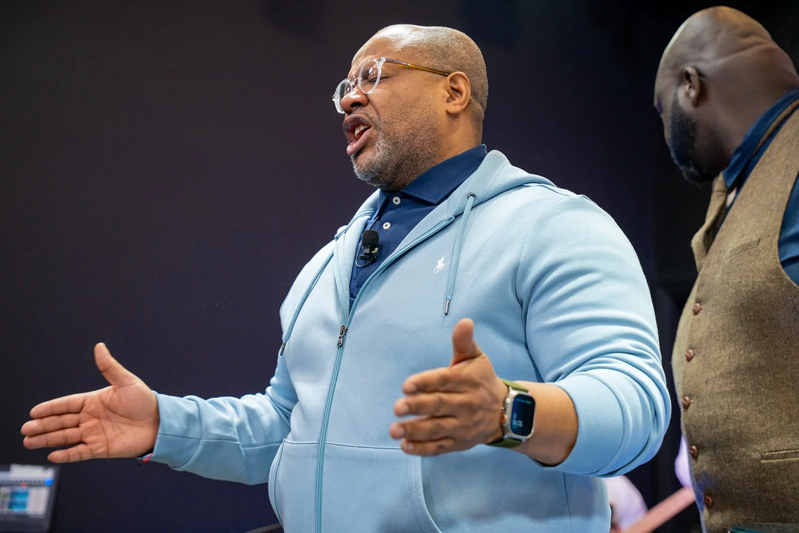 A man with glasses and a beard wearing a light blue hoodie and a polo shirt is speaking and gesturing with his hands. Another man is standing nearby, wearing a brown vest and a blue shirt, listening.