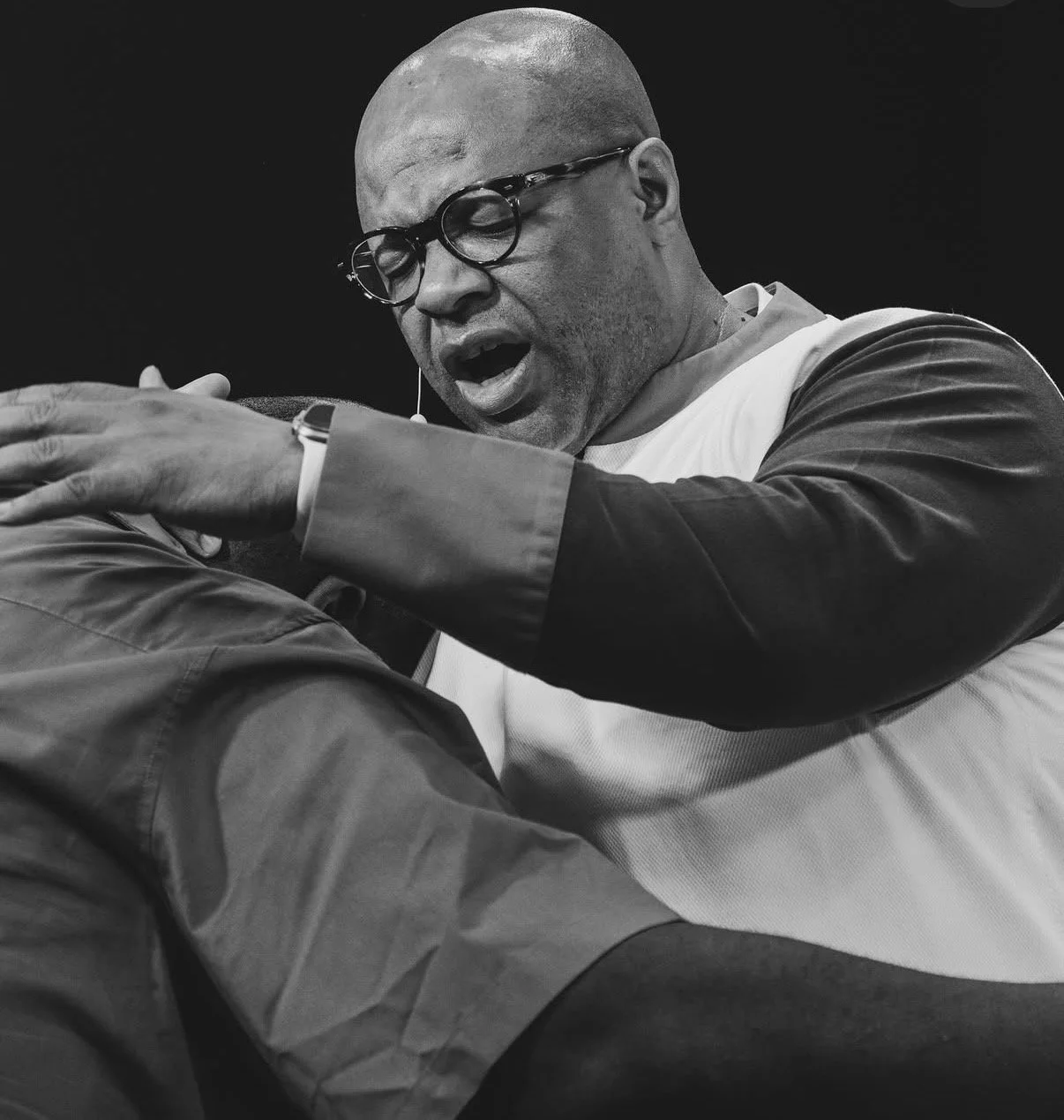 A black-and-white photo of a man with glasses, bald head, and facial hair, speaking passionately with his eyes closed and one arm extended outward.