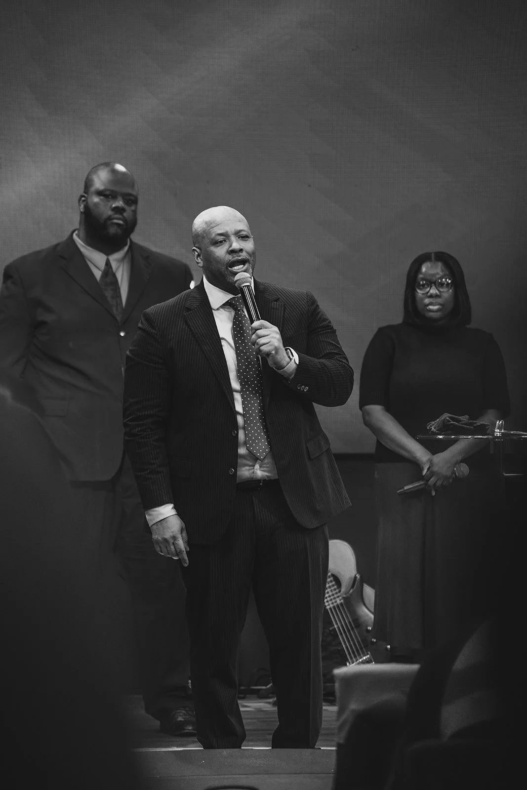 Black and white photo of three people on stage, with a man holding a microphone and speaking, accompanied by a woman holding papers and a man standing behind him, in a formal setting.