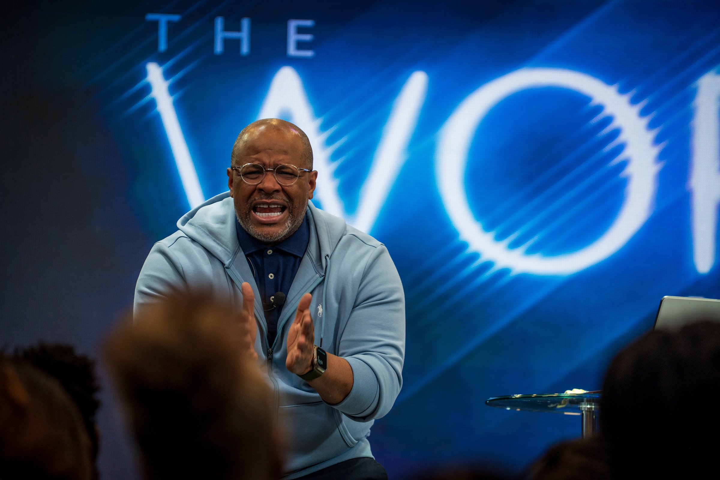 A man wearing glasses and a light gray hoodie appears to be speaking passionately during a presentation or discussion, with a large logo or graphic in the background.