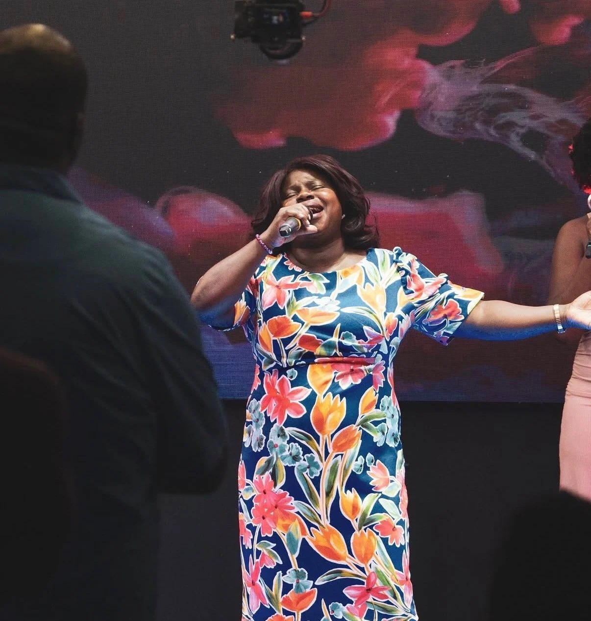 A woman singing passionately into a microphone on stage, wearing a colorful floral dress. There are people around her, including a man in a dark shirt and a woman on the right.