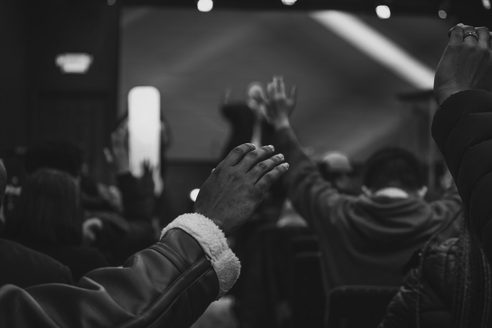 People seated in a dark room, some with hands raised, possibly in prayer or participation in an event, with a large screen in the background.