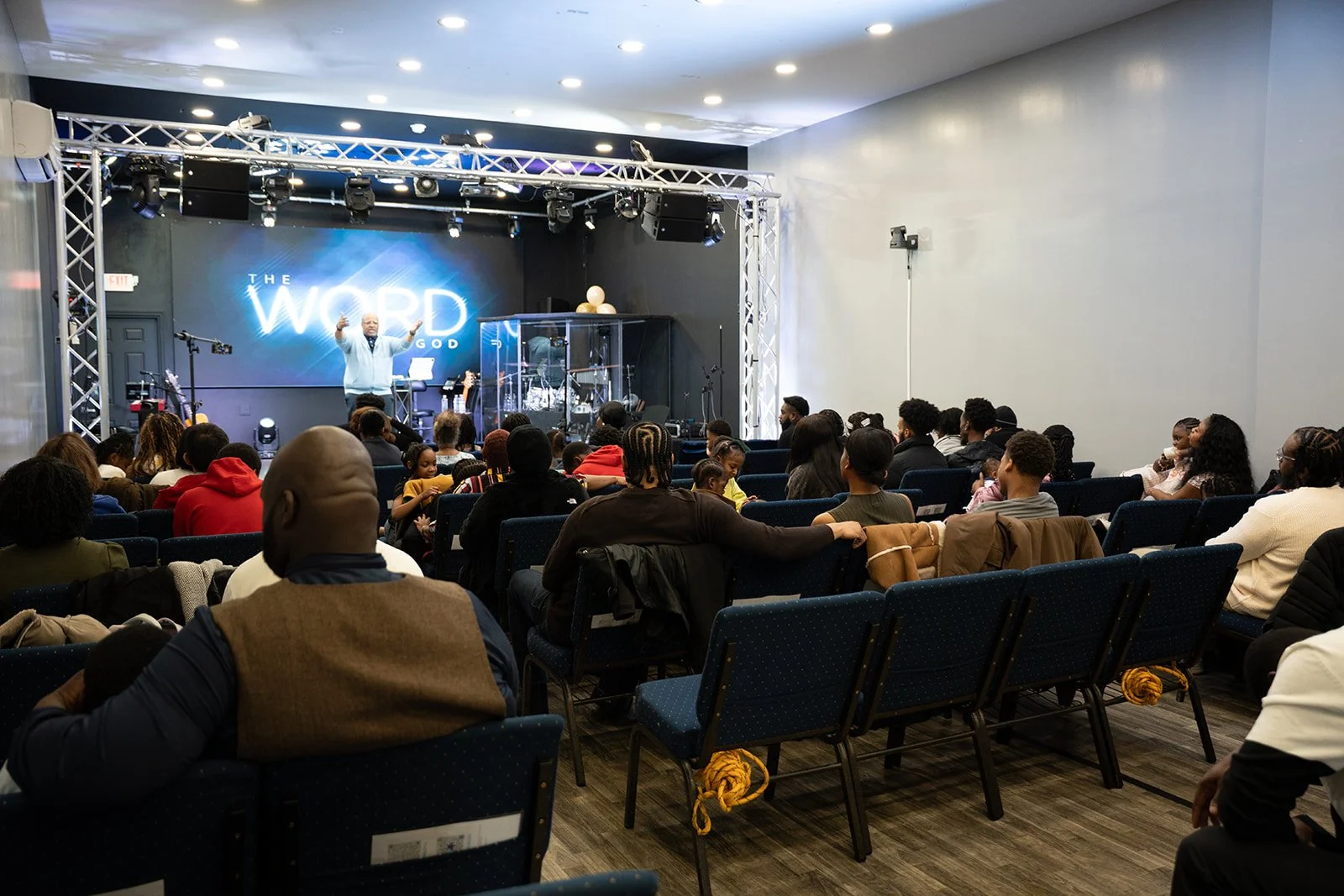A man speaking on stage during a religious service at a church or conference, with an audience seated in chairs facing the stage, and a large digital screen in the background displaying the words 'THE WORD OF GOD'.