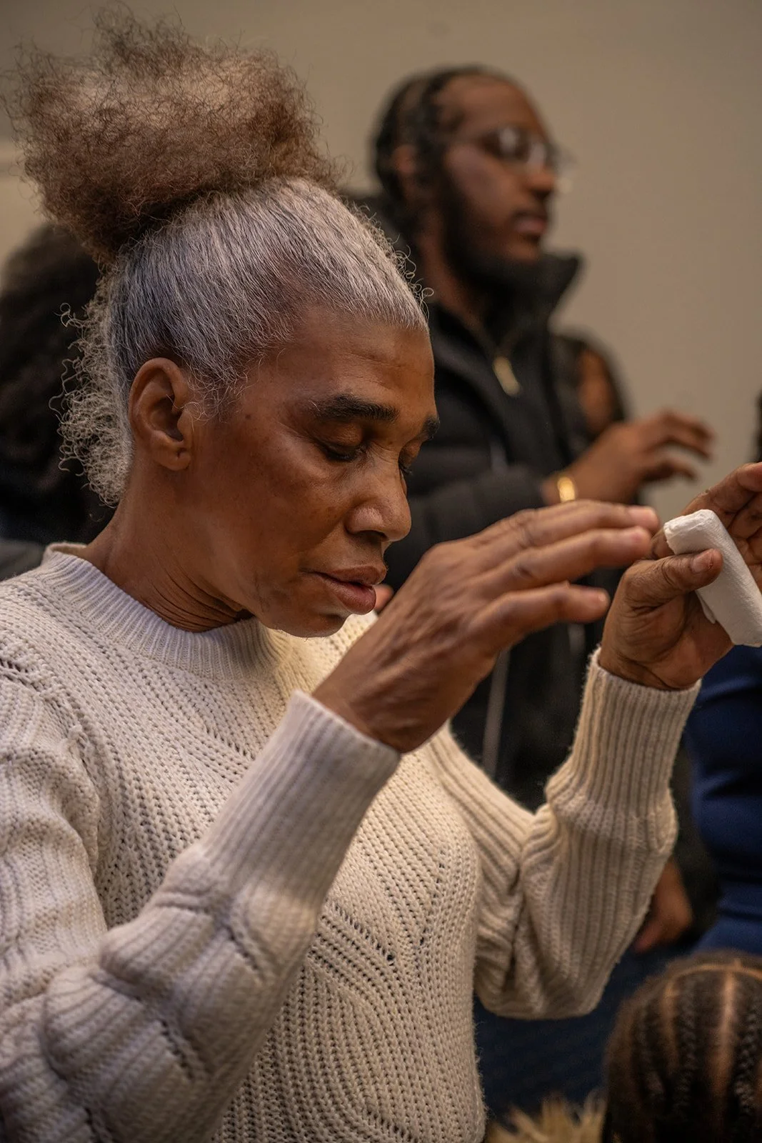 An elderly woman with gray hair tied up, wearing a cream-colored knitted sweater, holding a tissue or cloth to her nose, with eyes closed, in a somber moment, with two other women in the background, one with glasses and braided hair.