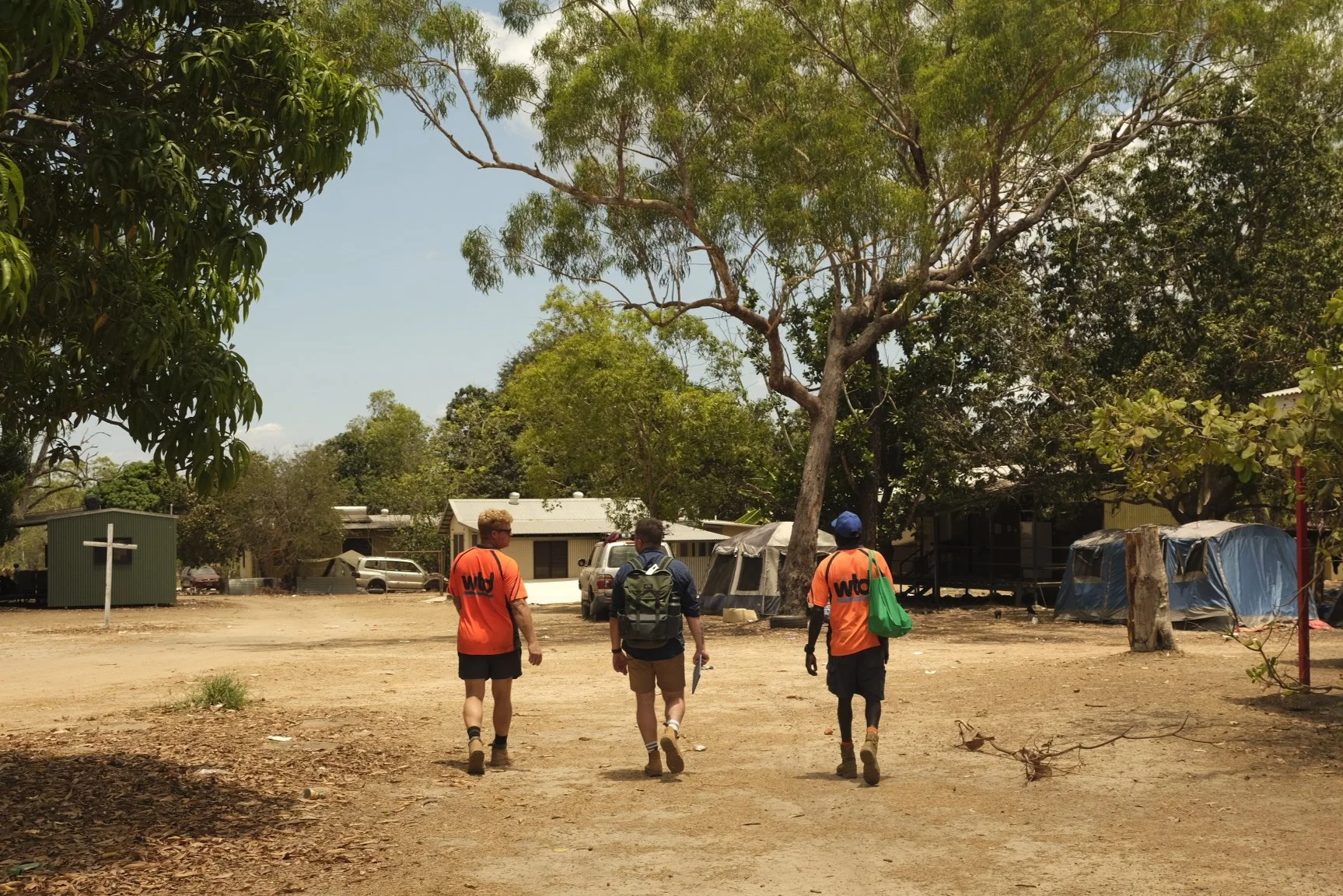 Three people walking in an outdoor area with trees, tents, and parked vehicles in the background.