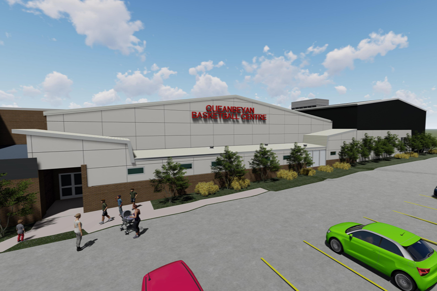 A large sports arena named Queanbeyan Basketball Centre with white and black exterior walls, a parking lot with green and pink cars, and a few people walking outside on a sunny day with blue sky and scattered clouds.