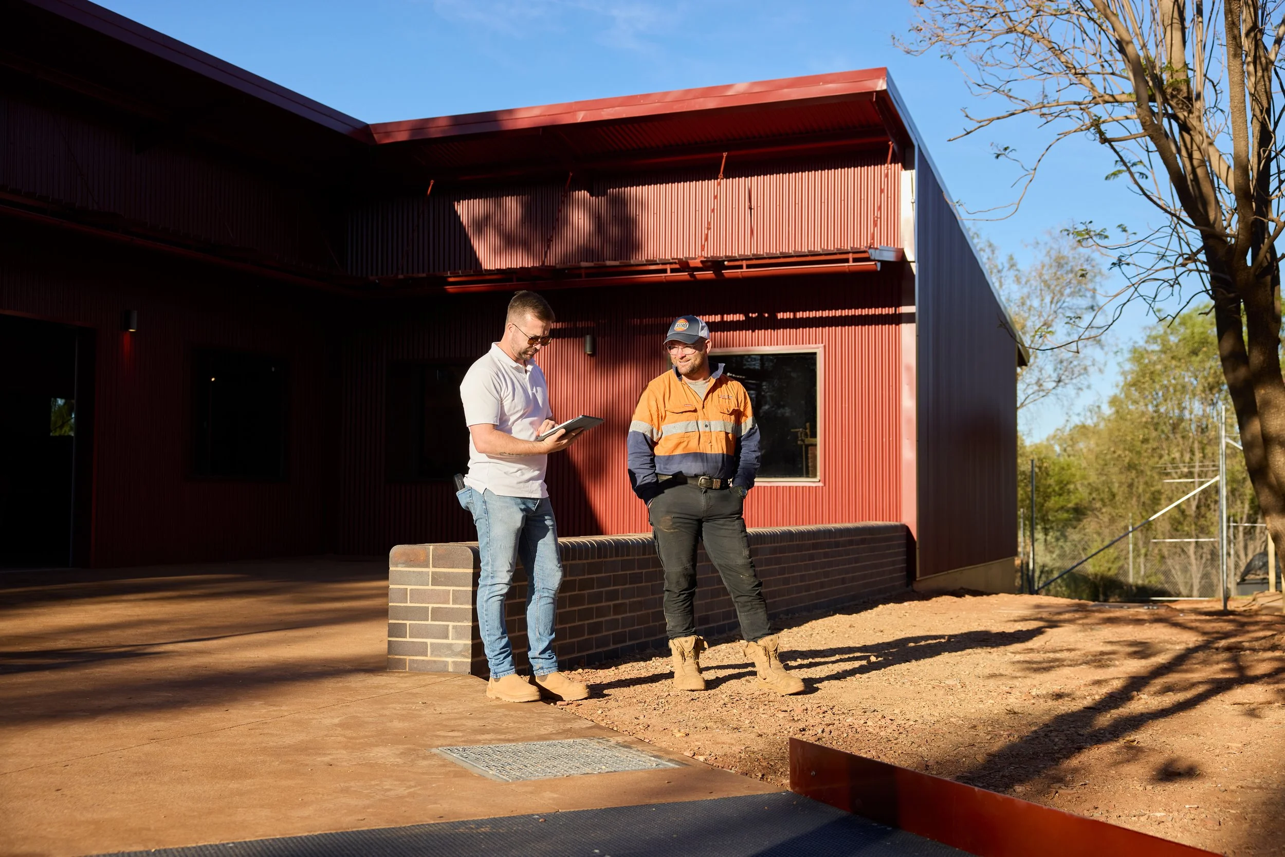 Two men stand outside a red building, one holding a tablet and the other in work boots and a high-visibility jacket, on a dirt lot during sunny weather.