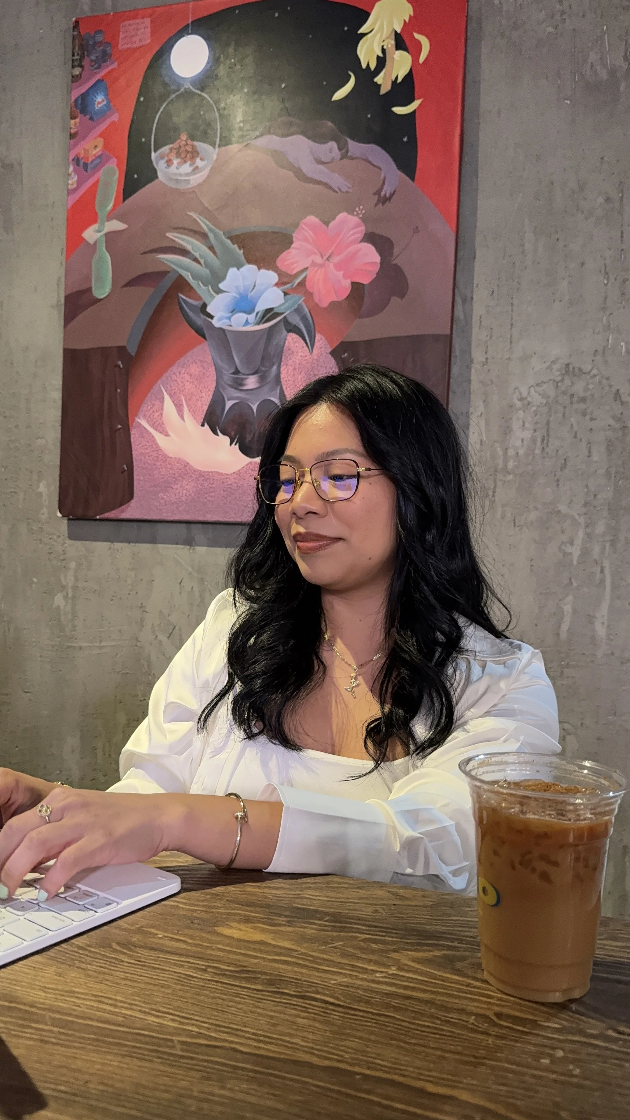 Woman with long black wavy hair and glasses working on a white keyboard at a wooden table, wearing a white top and gold jewelry, with iced coffee, in front of a colorful abstract painting in a modern workspace.