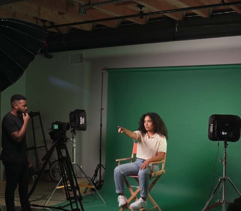 Video director guiding a shoot in a green screen studio, with professional lighting and camera equipment, while an assistant observes.