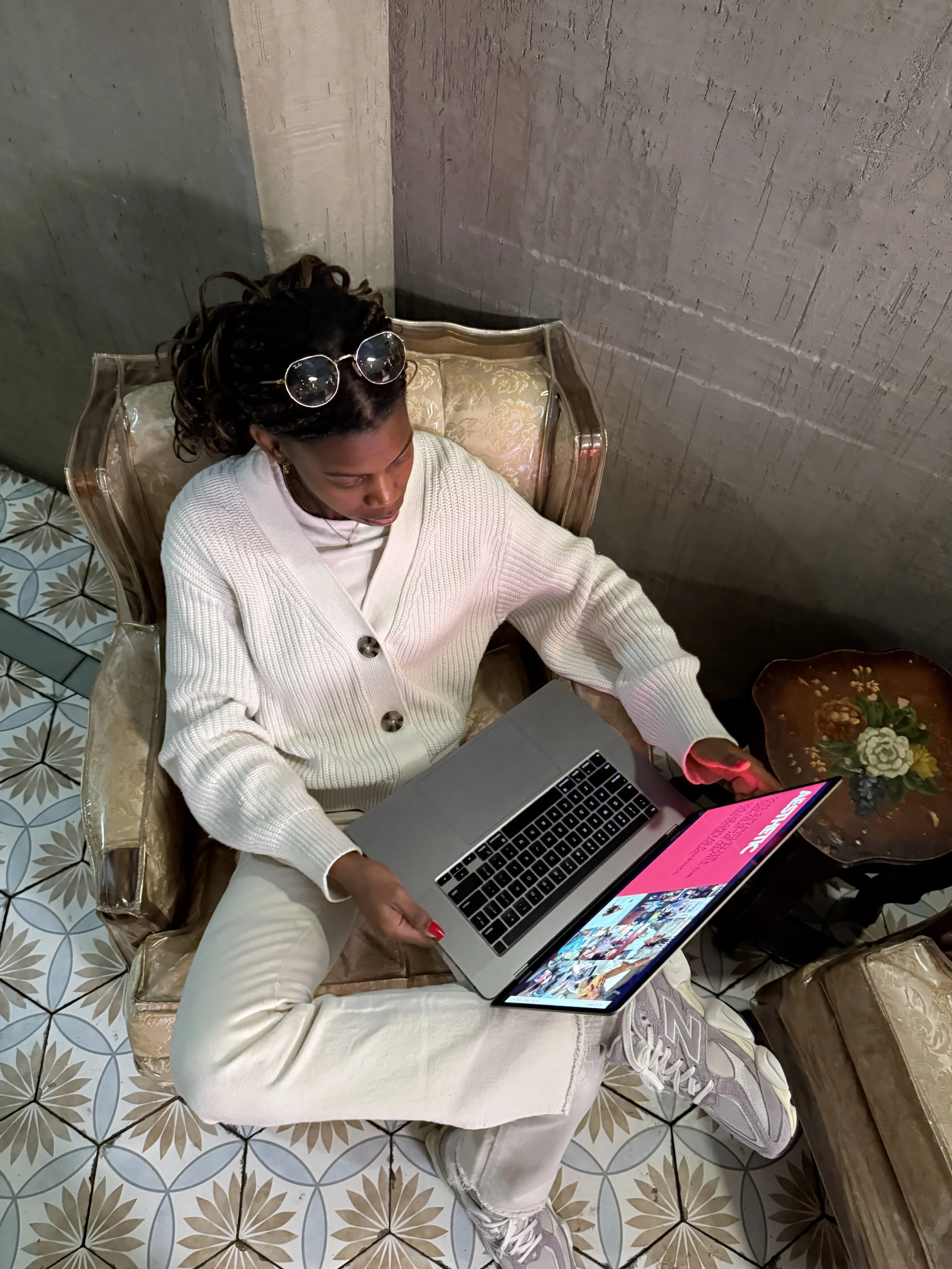 Woman sitting in a vintage armchair using a laptop, wearing a white cardigan, white sneakers, and sunglasses on her head, next to a small table with flowers, in a room with patterned tile flooring and a concrete wall.