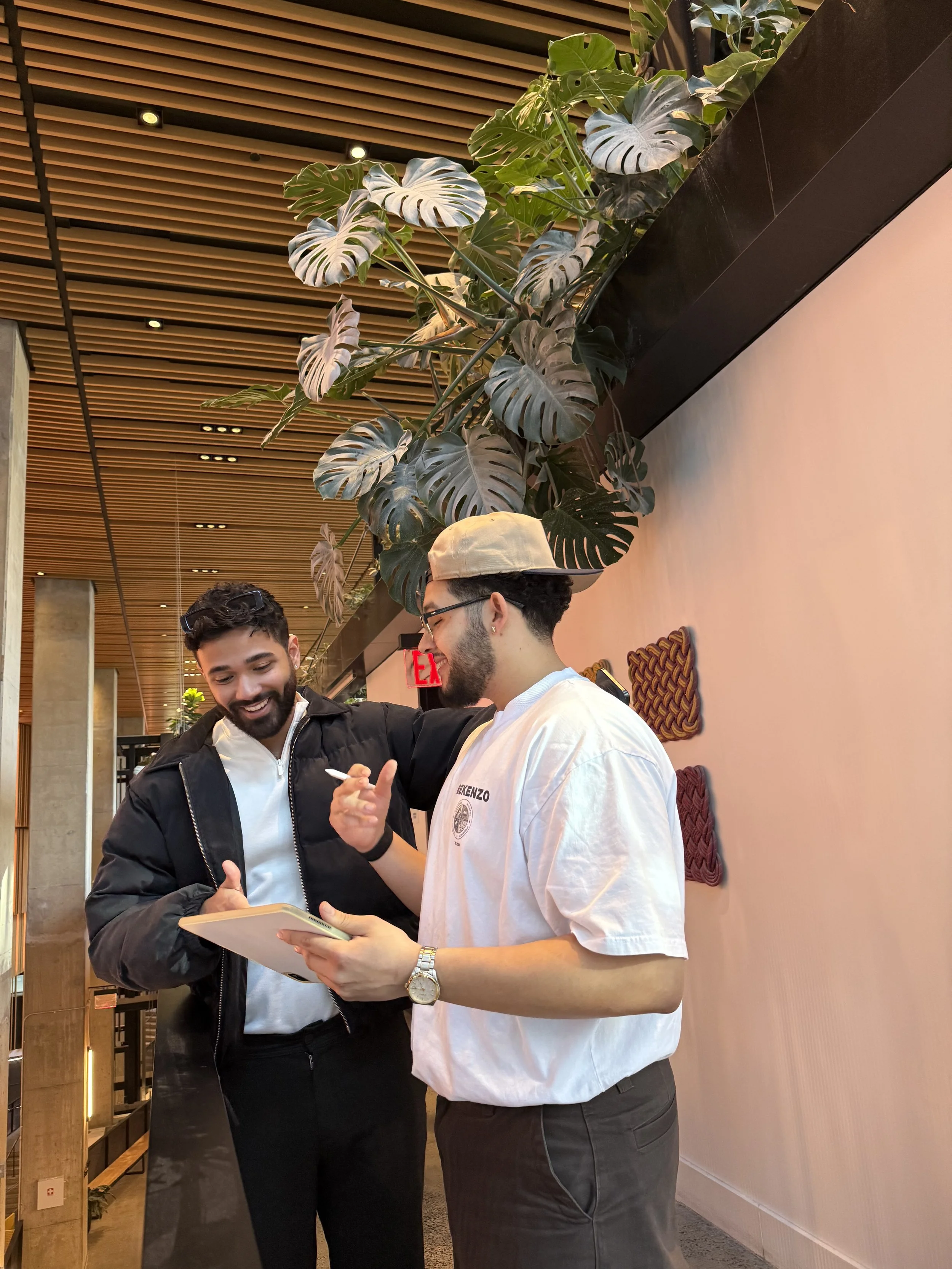 Two men smiling and reviewing notes on a notepad, one holding a pen, collaborating in a modern indoor workspace with plant decor.