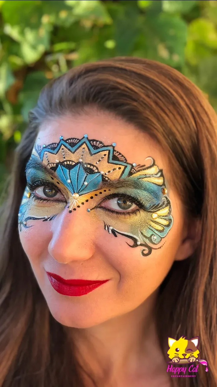 A woman with detailed butterfly face paint in blue, yellow, and black, with decorative dots and swirl accents, wearing bright red lipstick, outdoors with green foliage in the background.