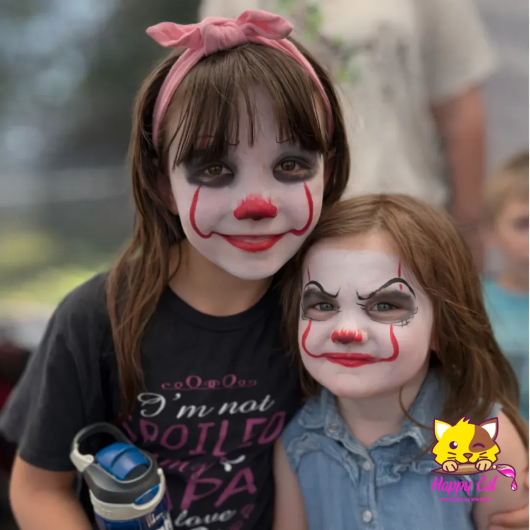 Two young girls dressed as clowns with white face paint, red noses, and black and red makeup, standing close together outdoors. One girl has a pink headband and brown hair, and the other has red hair and a denim shirt.
