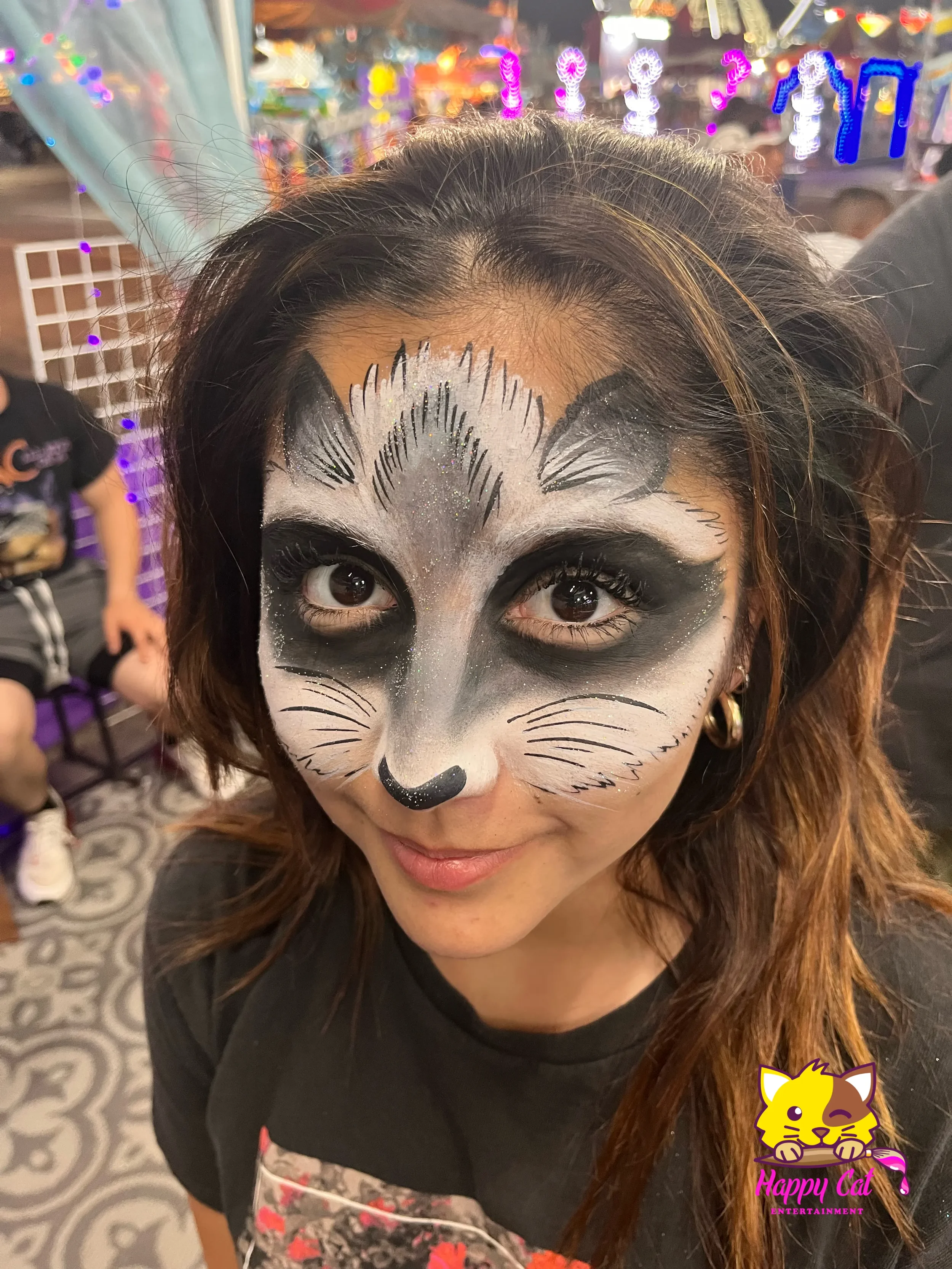A woman with face paint resembling a cat, with black and white colors, glitter, and detailed whiskers. She is smiling at the camera in an amusement park with colorful lights in the background.