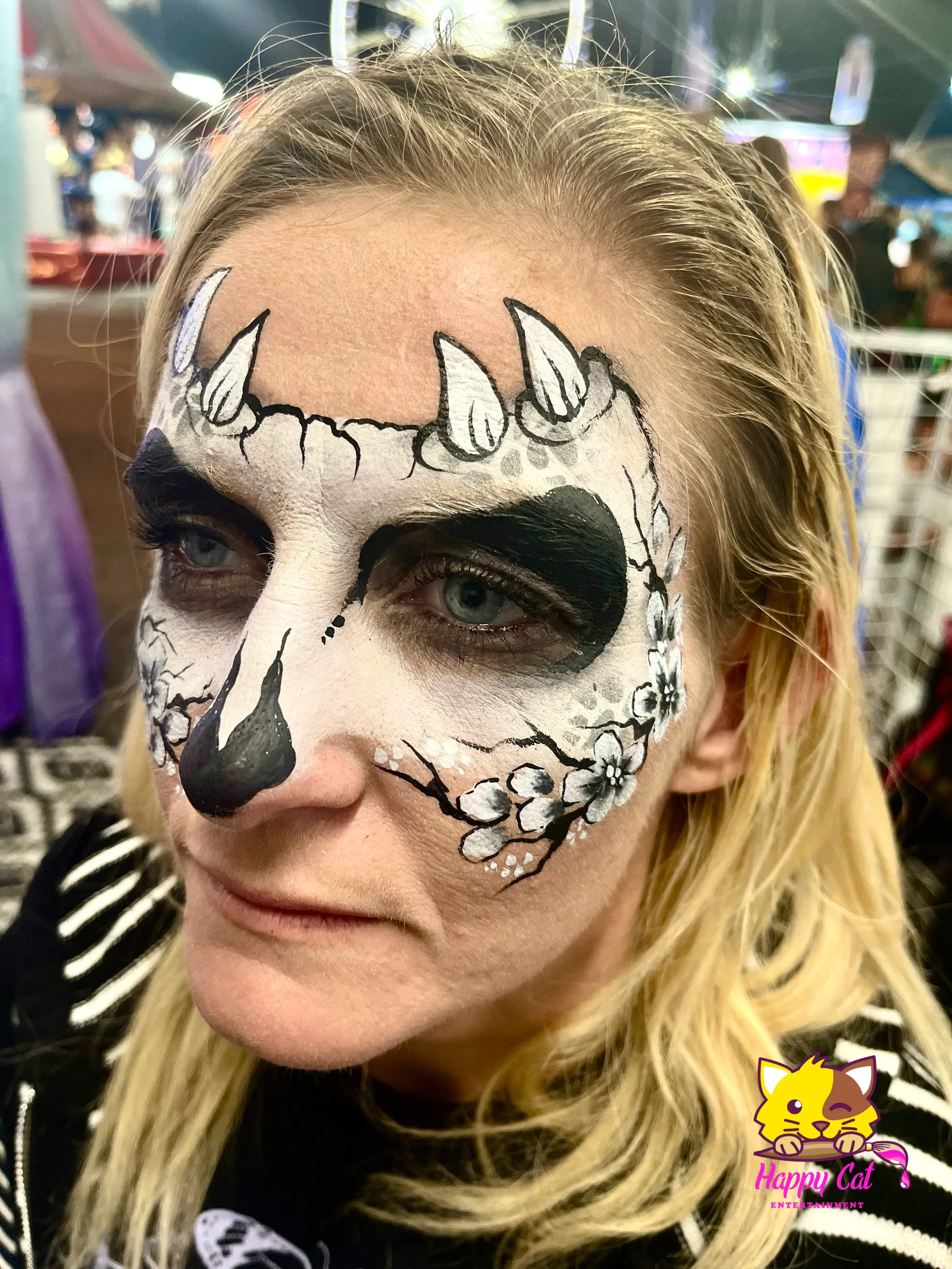 A woman with Day of the Dead face paint, featuring a white base, black around the eyes, nose, and mouth, and decorative black and white floral and line details, at a festive event.